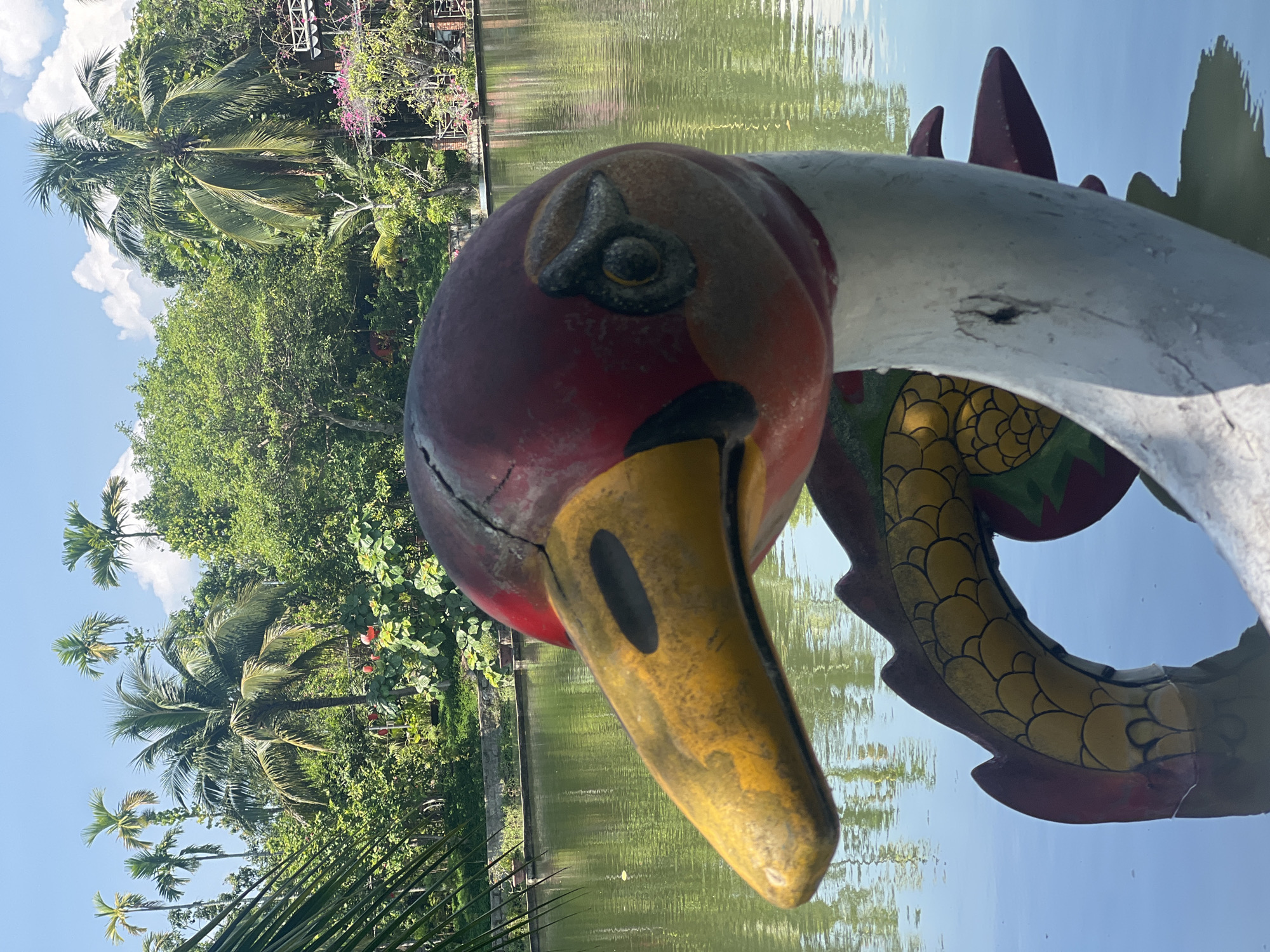 A brightly colored, weathered statue of a mythical creature, possibly a dragon or phoenix, floats in a tranquil body of water. The statue's head, predominantly red with a yellow beak and black eye details, is in the foreground, slightly off-center to the right. Its body, adorned with yellow scales and red accents, curves into the water. The background features a lush, green landscape with numerous palm trees, dense foliage, and a clear blue sky with scattered white clouds. The water reflects the surrounding greenery and sky. The scene is likely a public park or recreational area within the city of Suối Tân, Vietnam, given the characteristic tropical vegetation.