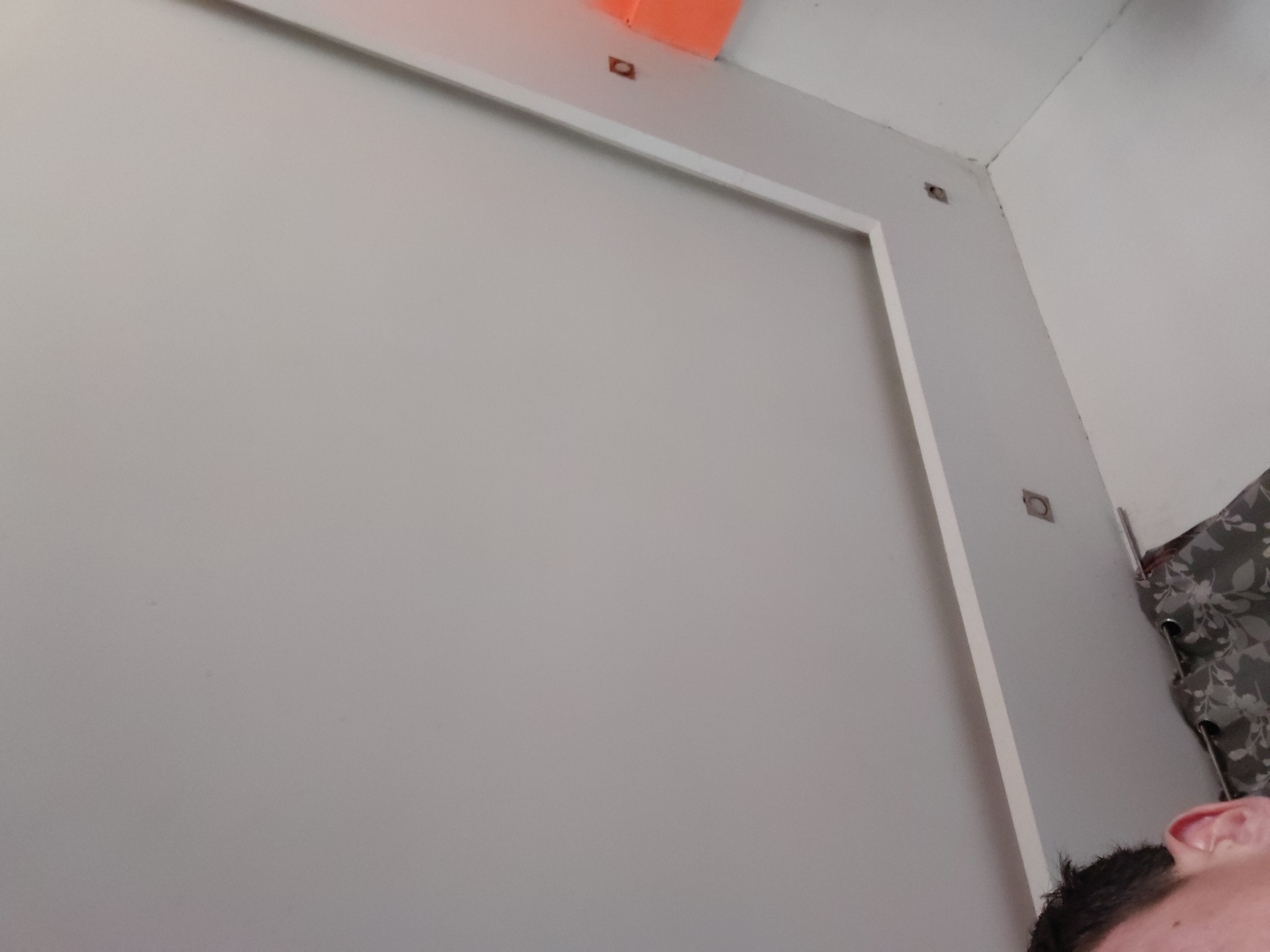 An upward-angled, diagonal shot captures the interior of a room, presumably in Santa Ana, Venezuela. The dominant features are light-colored (off-white or light grey) walls, one of which displays a prominent, large white rectangular frame or panel. This frame runs vertically and horizontally, bordering a central area of the wall.
Along the top edge of the main wall, above the white frame, several small, dark, square or rectangular elements are embedded, appearing to be recessed fixtures or mounting points. One of these is situated beneath a bright orange, possibly plastic or painted, rectangular object or surface that protrudes slightly from the wall near the ceiling line on the upper left. Another similar embedded element is visible further to the right, near the ceiling corner, and a third is visible on the right vertical wall.
In the bottom right corner of the frame, a partial view of a person is visible, showing the top of their head with dark hair and their right ear. To the far right, a section of a patterned curtain or fabric, featuring a floral or leaf design in shades of grey and white with dark eyelets, hangs near a corner of the room. The overall impression is of a modern, clean indoor space. No text is visible in the image.