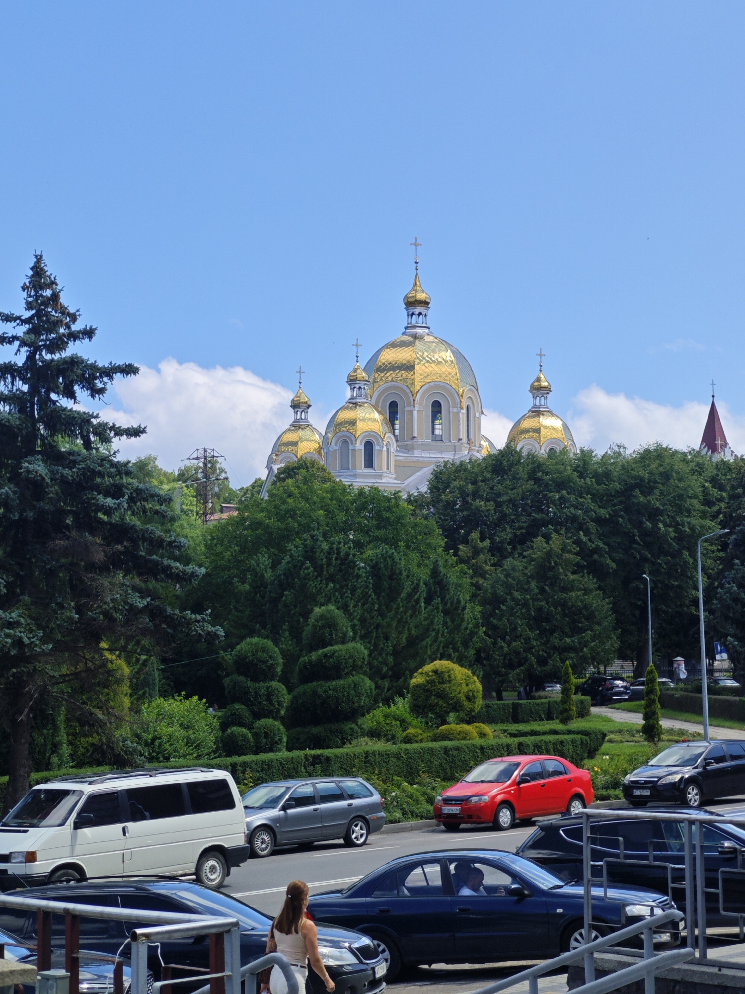 A daytime, exterior, medium shot captures a street scene in front of a large, ornate cathedral with golden domes. The cathedral is partially obscured by lush green trees and manicured bushes. Several cars are visible on the road, including a white van, a gray station wagon, a red sedan, a black SUV, and a dark-colored sedan with a driver. A woman with long brown hair is standing by the side of the road. The sky is blue with scattered white clouds. The overall impression is a sunny day in a city with a prominent religious building.