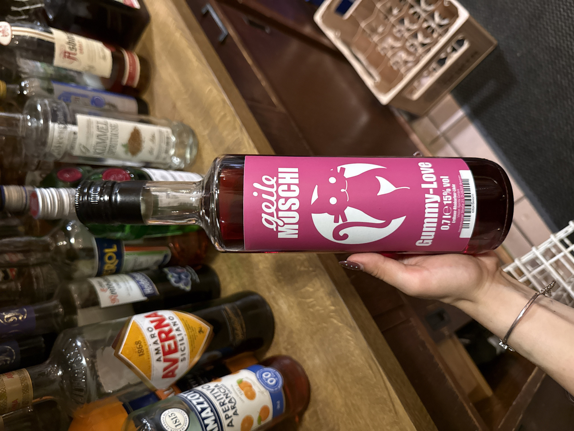 The image shows a person holding a bottle of "Gummy-Love" liquor with a pink label featuring a cartoon cat. The label also includes "geile MUSCHI" text. The bottle is surrounded by other liquor bottles in what appears to be a bar or storage area. Several brands such as "Averna", "Mazzotto", and "Kümmel" are partially visible. The liquor is produced by Vision Spirits GmbH, and the bottle states they are located at Neudenburger Straße 21, D-28207 Bremen, Germany. No location of Bruchköbel, Germany is mentioned on the bottle. The person holding the bottle has a bracelet and manicured nails.