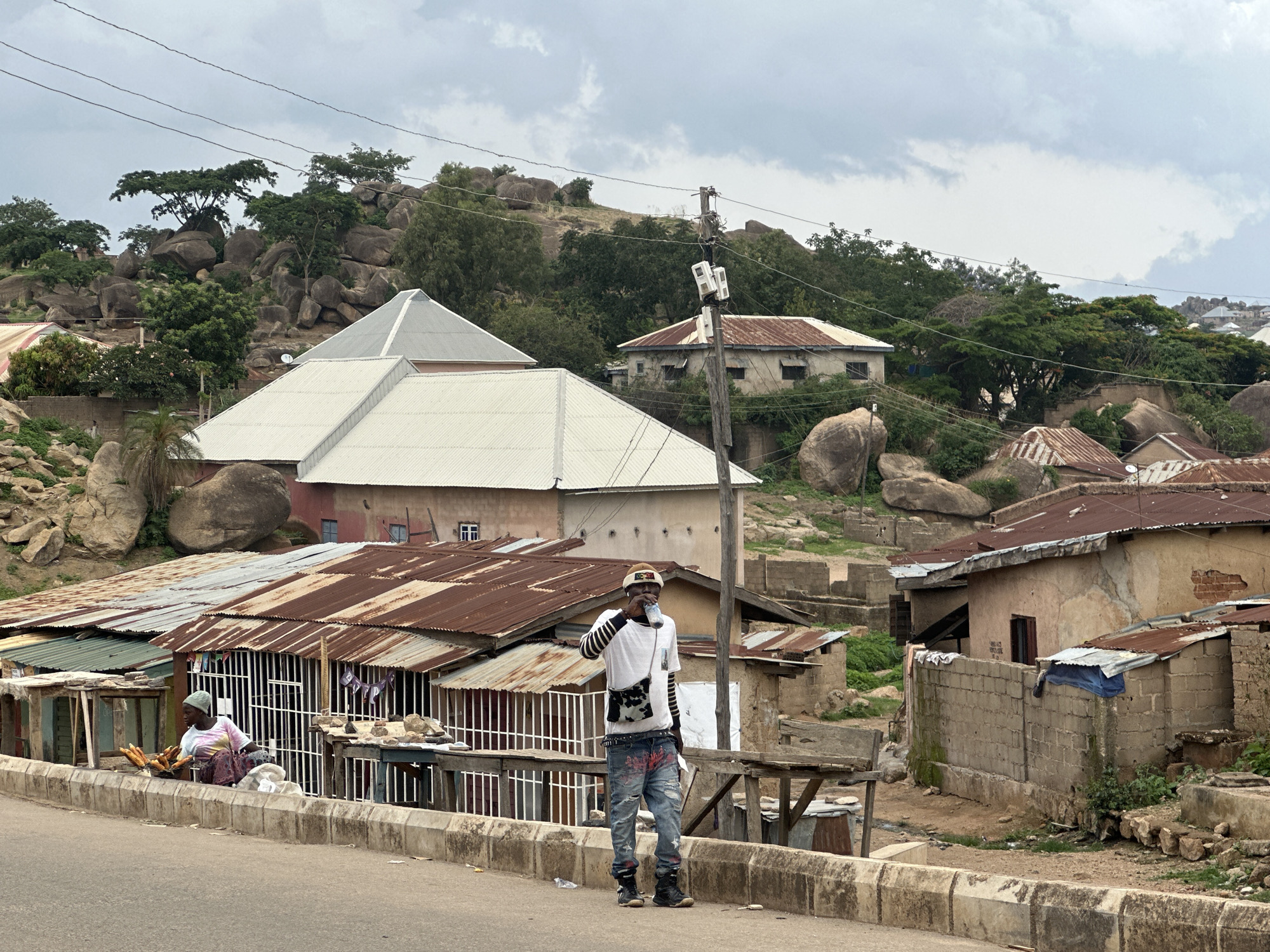 The image depicts a street scene in Jos, Nigeria. In the foreground, a man stands on a low concrete barrier adjacent to a road, wearing jeans, a white t-shirt, a hat, and a face mask. He appears to be drinking from a container. Beside him, a woman sits on a stool next to a metal structure, possibly selling food, with corn on display. The backdrop features densely packed, low-rise buildings with corrugated metal roofs. A large, light-colored building with a distinctive peaked roof is visible behind the street vendors. A hill with large rocks and trees rises in the background. An overhead power line crosses the upper part of the image. The sky is overcast.