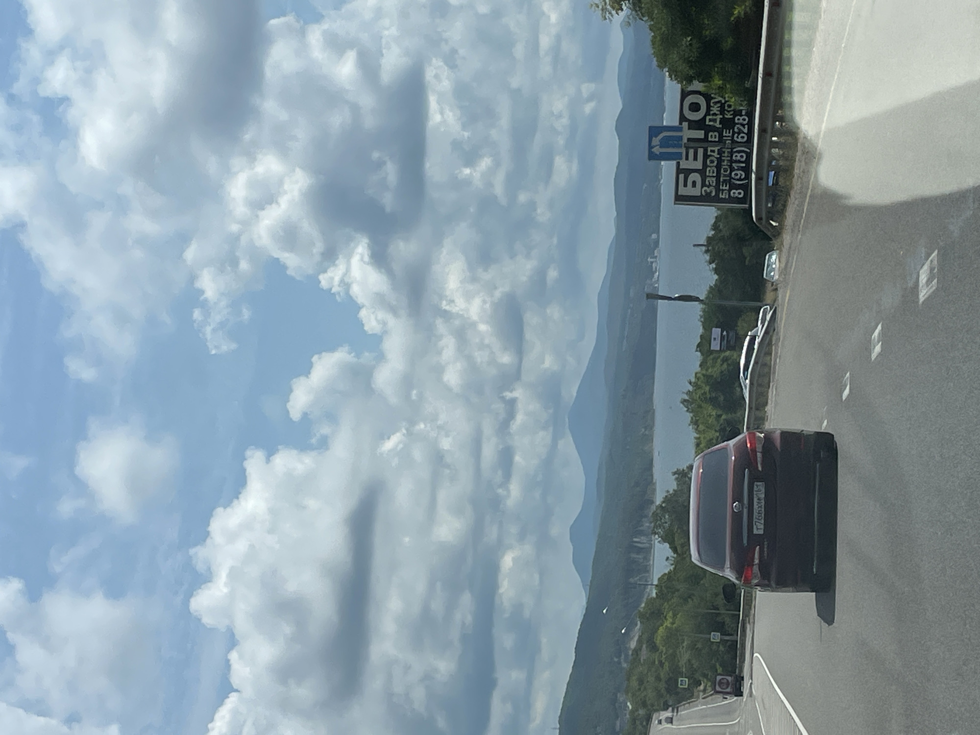 A car is driving on a highway in Dzhubga, Russia. The car is a dark red sedan with a license plate that reads "T766XA 161". The highway is paved and has white lane markings. There are mountains in the background and a body of water. There is a billboard on the right side of the road that says "БЕТОН Завод в Джу" which translates to "CONCRETE Factory in Dzhubga". The billboard also includes a phone number. The sky is cloudy.