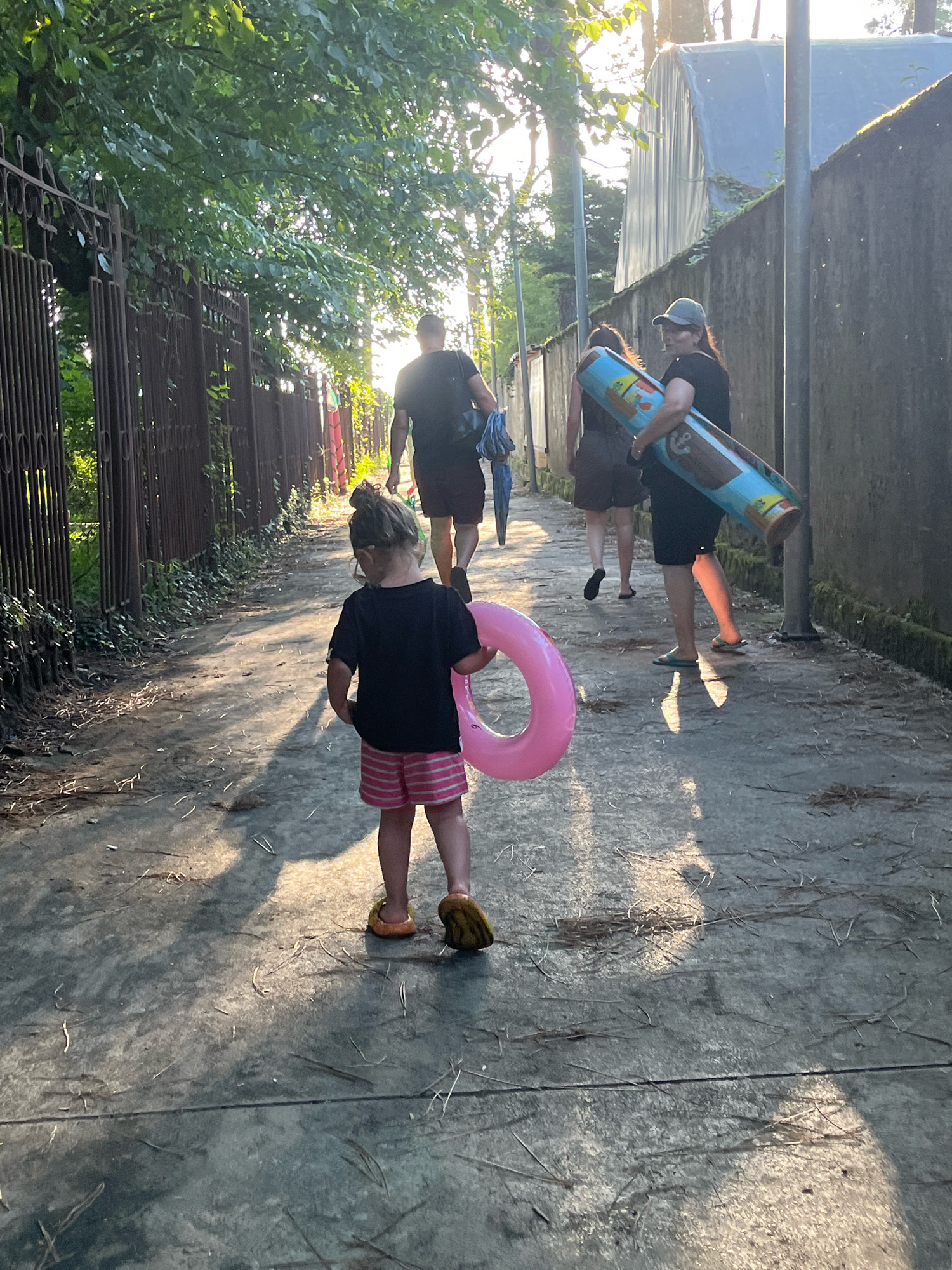 The image shows a group of people walking down a paved path in Ureki, Georgia, towards a body of water. The path is bordered by a rusted metal fence on the left and a concrete wall on the right. Lush green trees with dappled sunlight filter through the leaves, casting shadows on the path. In the foreground, a young child, seen from behind, wears a black t-shirt and pink striped shorts, holding a pink inflatable ring. Behind the child, two adults are walking. One adult is carrying a large, blue inflatable tube with colorful cartoon images. The other adult, partially visible, is wearing a baseball cap and dark clothing. The lighting suggests it is late afternoon or early evening, with the sun casting long shadows. The overall atmosphere is one of a casual outing, possibly to the beach, given the presence of inflatable water toys.