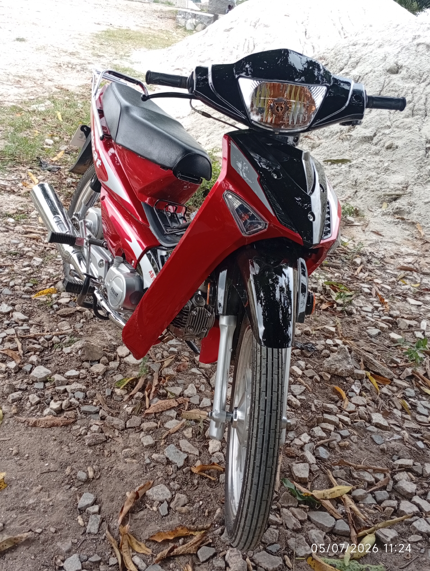 A red and black motorcycle is parked on a gravelly path, possibly in Bana Stream, Nigeria. The motorcycle is facing towards the viewer and is angled slightly to the right. Its front wheel is visible, as is part of the engine and exhaust pipe. The motorcycle has a black seat and handlebars. In the background, there is a pile of light-colored sand and some dry leaves scattered on the ground. A timestamp in the bottom right corner reads "05/07/2026 11:24".