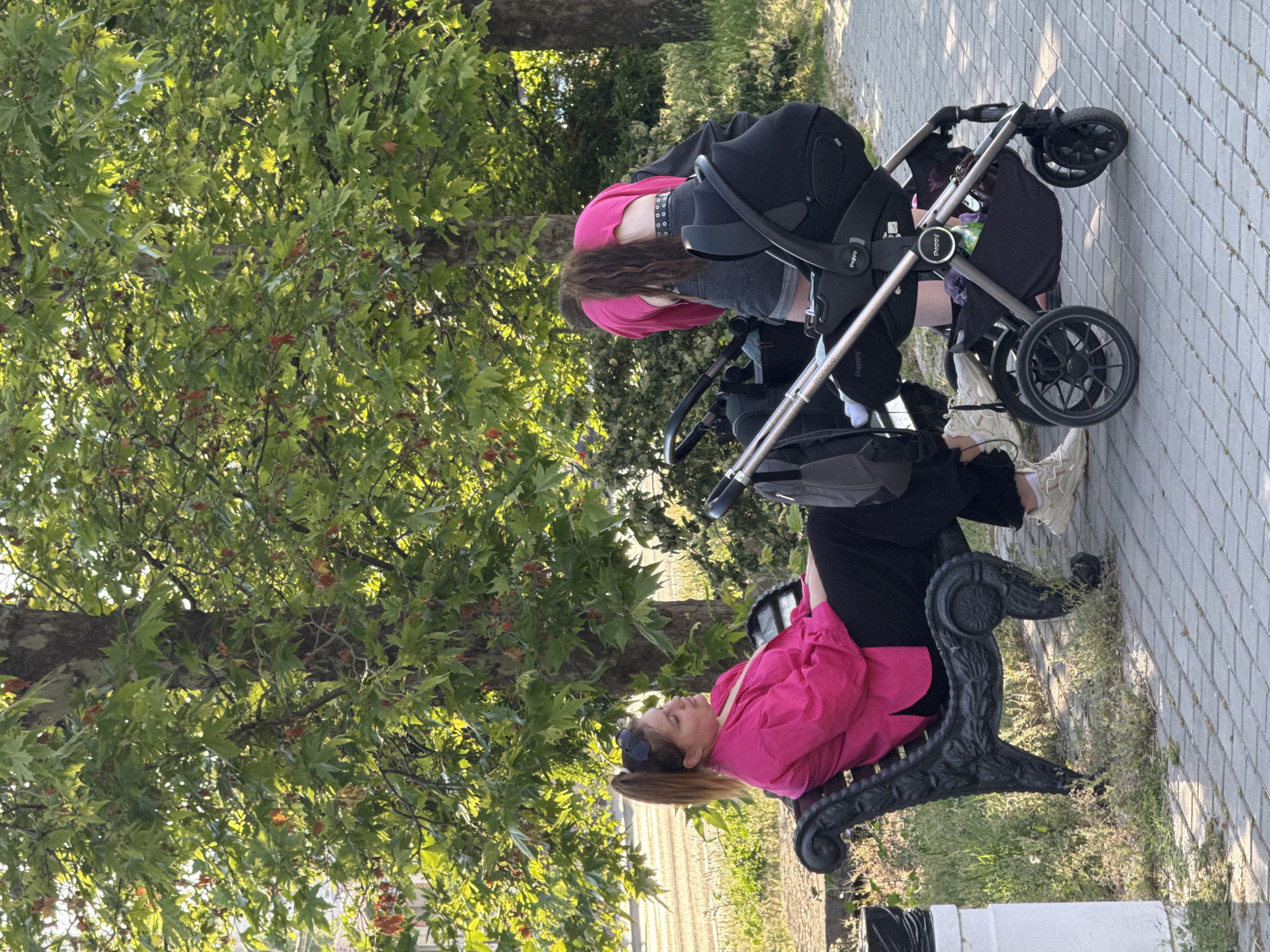 In a park setting, two women and a baby stroller are visible. One woman, wearing a bright pink shirt and black pants, is seated on a decorative black bench. She has brown hair pulled back in a ponytail and is wearing sunglasses on her head. She is looking to her left, with her arms resting on the bench. The second woman, wearing a pink crop top and denim shorts, is bent over, attending to the baby stroller. Her long brown hair is down her back. The stroller is black with silver accents and a black infant car seat is attached. The ground is paved with light-colored interlocking bricks, and patches of green grass and weeds are visible around the bench and pavement. Large trees with green leaves and some reddish-brown seed pods dominate the background, casting shadows. Sunlight filters through the leaves. A white trash receptacle is partially visible on the far left of the image.