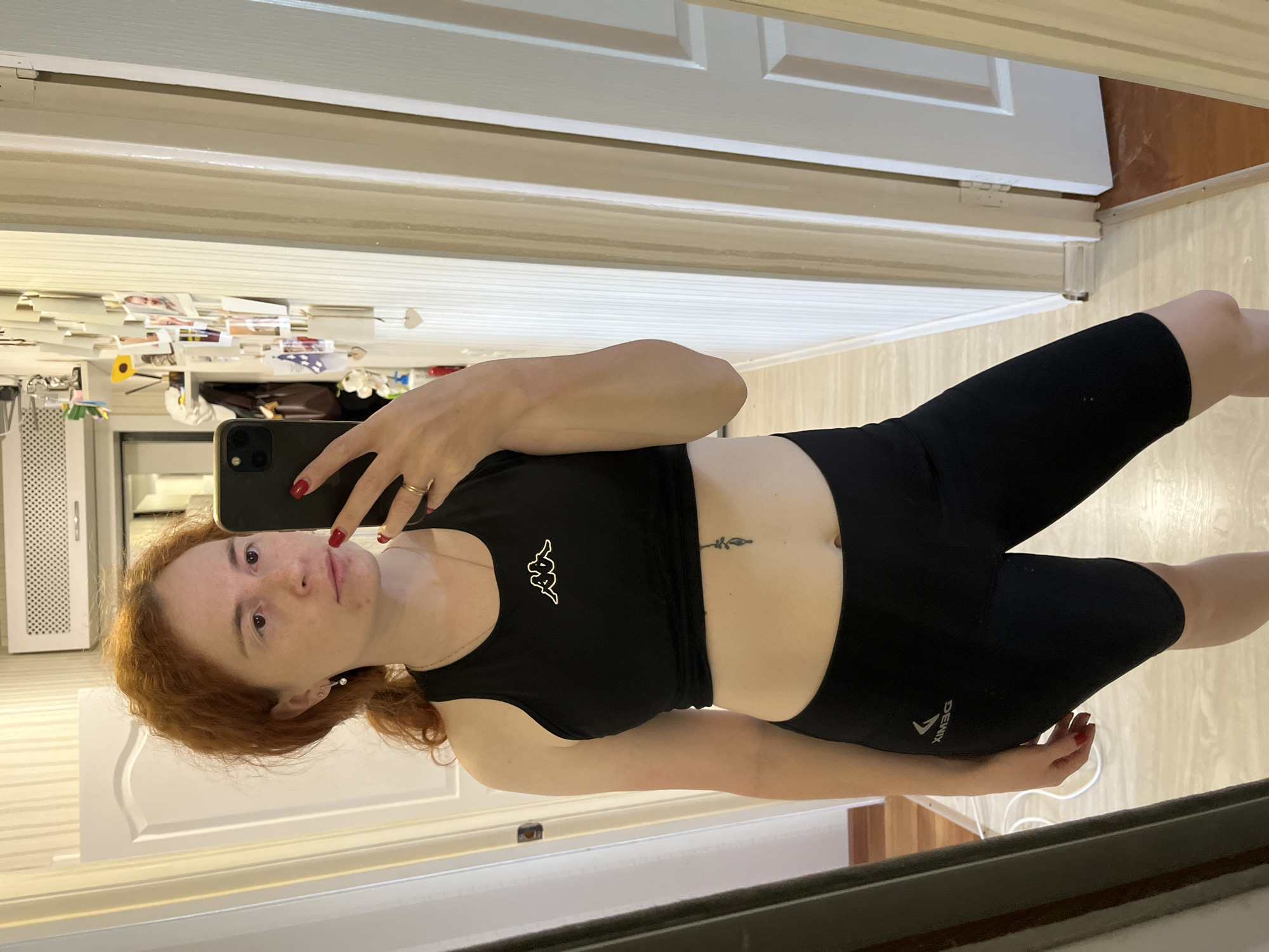 A red-haired woman is taking a selfie in a mirror. She is wearing a black Kappa crop top and black Dewix biker shorts. The background includes a refrigerator covered in photos and papers, a white door, and a hallway with light-colored walls. Based on the user's location, this is likely in Chaikovsky, Russia.
