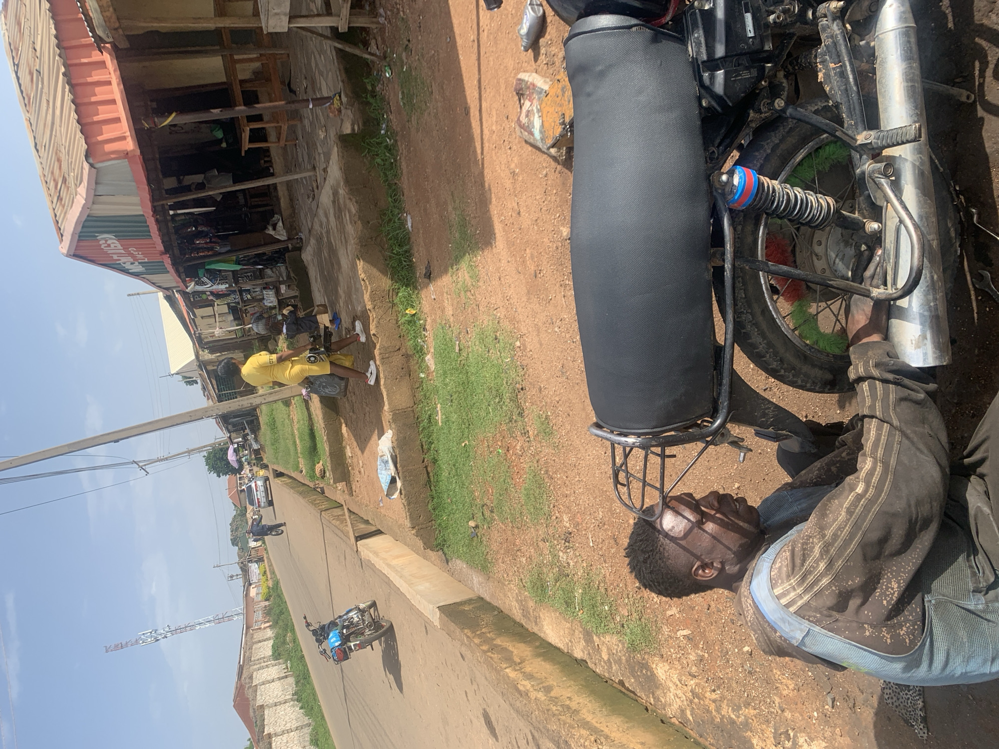 The image depicts a street scene in Gura Top, Nigeria, on a sunny day. In the foreground, a man wearing a dirty blue and brown mechanic's jumpsuit is working on the rear wheel of a black motorcycle, which is positioned on the dusty ground. The motorcycle has a prominent exhaust pipe and a shock absorber with red and blue bands. Behind the man and the motorcycle, a paved road stretches into the distance, lined with a concrete drainage channel.
On the roadside, a woman in a yellow outfit walks away from the camera, carrying a large black plastic bag. Several shops with corrugated metal roofs line the right side of the street. One shop's facade displays the text "NWSGI LUGBO". Various items are visible for sale or storage within the shops and outside. Further down the road, a blue motorcycle is parked on the left side, and a white car is driving away. In the background, a tall cellular tower with white and red markings is visible against a pale blue sky. The environment appears to be a busy urban or semi-urban area, with evidence of commerce and vehicular traffic.