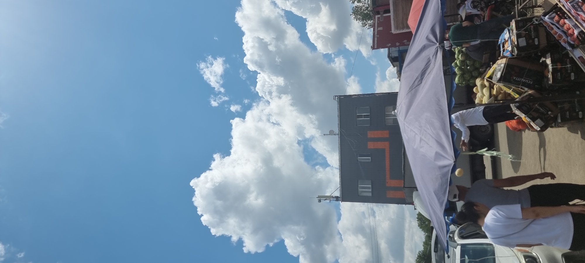 An outdoor market scene unfolds under a bright blue sky with scattered white clouds in Aktobe, Kazakhstan. In the mid-ground, a dark gray building with an orange geometric design on its facade stands tall, topped with two communication antennas and satellite dishes. The building has several rectangular windows on its upper floors. In the foreground, a large, light purple market umbrella provides shade over a produce stall. Beneath the umbrella, a vendor in a white shirt appears to be tossing a yellow melon into the air, while a person in a gray shirt and white cap stands nearby, facing away from the camera. Further to the right, a stack of watermelons and crates of fruit, including peaches, are visible. Several vehicles, including a white car, are parked in the background, and a few other individuals are partially visible, suggesting a bustling marketplace. The environment is characterized by a mix of urban architecture and open-air commerce.