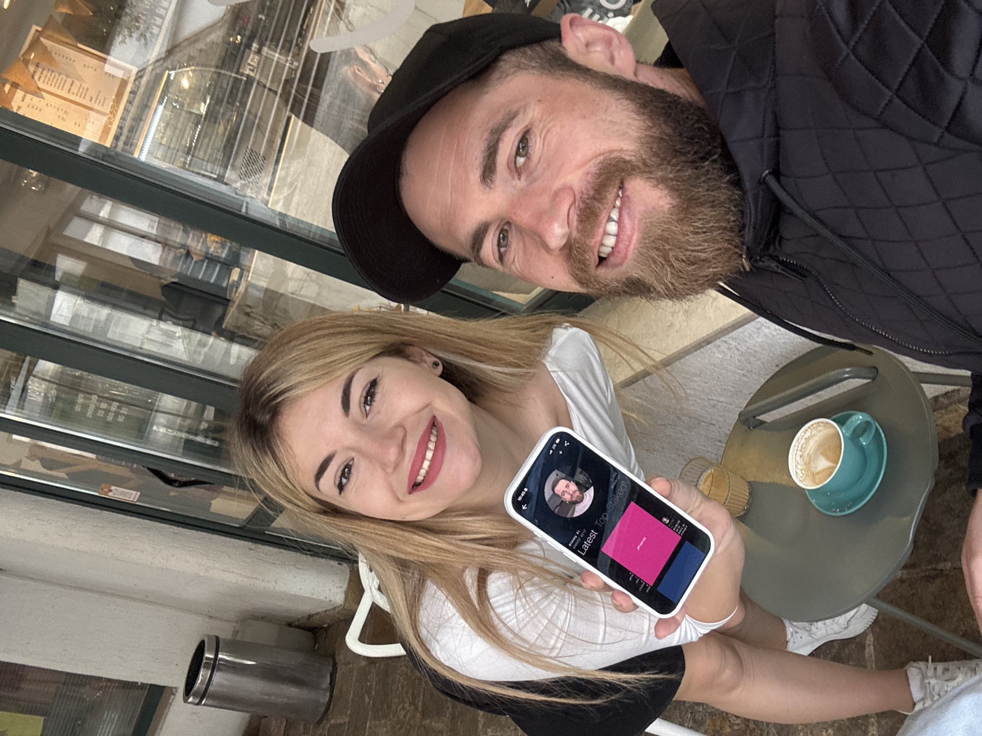 Two individuals, a male and a female, are captured in a selfie-style photograph within an indoor cafe setting. The male, positioned at the top right of the frame, wears a black baseball cap and a black quilted jacket. He has a beard and a smile. The female is positioned lower in the frame, holding a smartphone that displays a profile picture of the male. She also smiles towards the camera. On a round table next to them, there is a green coffee cup with some liquid inside and a small glass. A metal trash can is visible near a window, revealing an urban scene that suggests the location is likely within a city like Budva, Montenegro.