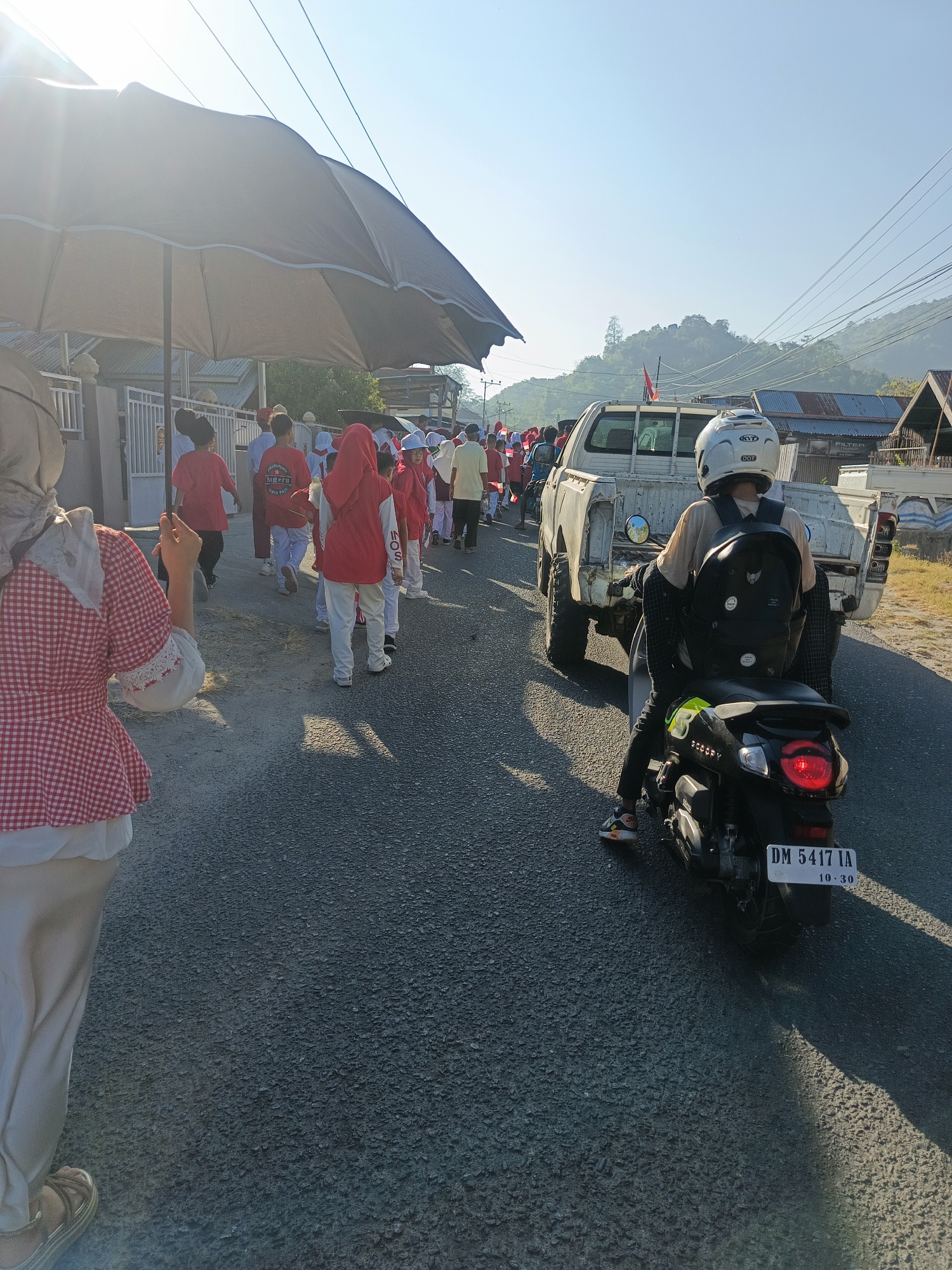In an outdoor street scene, likely within the city of Gorontalo, Indonesia, under a clear blue sky with soft sunlight filtering through, a group of children dressed in red and white uniforms is walking in procession. A large, dark umbrella is held aloft by an unseen person, casting a wide shadow that partially obscures the children and the foreground. The procession moves along an asphalt road, with a white pickup truck and a black scooter with a rider in a helmet following closely behind. The environment features modest buildings and vegetation in the background, suggesting a residential area. The overall atmosphere appears to be one of a communal event or celebration, possibly a parade.