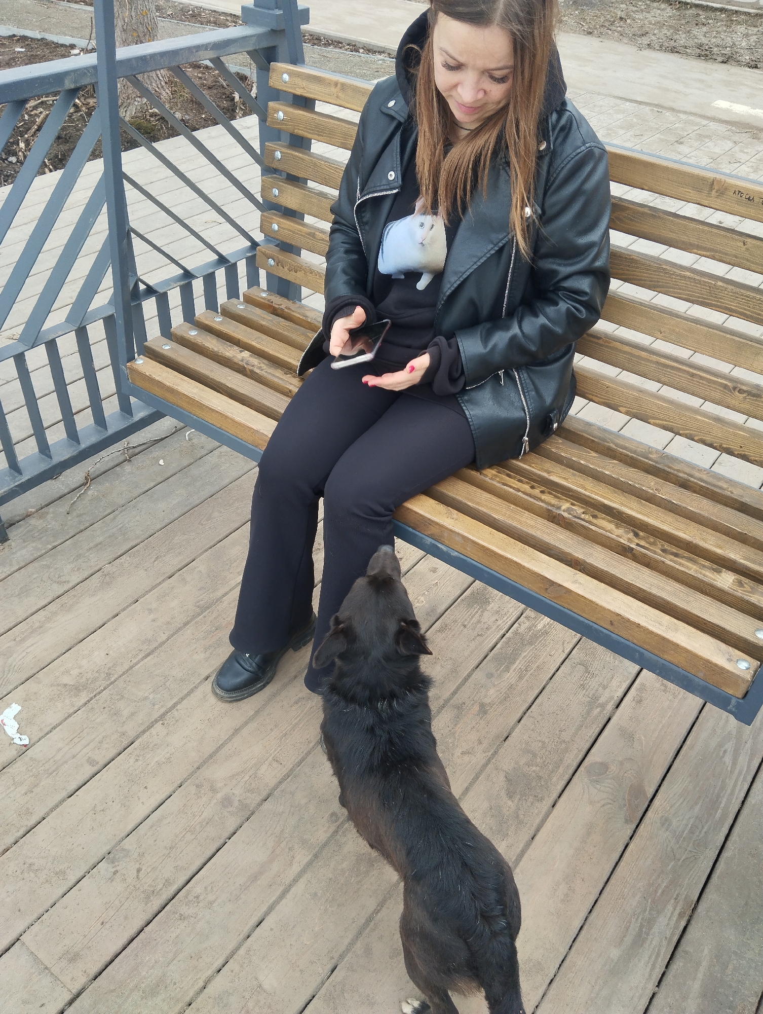 The image shows a young woman sitting on a park bench in Votkinsk, Russia. She has medium-length brown hair and wears a black hooded sweatshirt, a black leather jacket, and black pants. She is holding a smartphone and looking down at it. A black dog stands in front of her, looking up at her. The bench is made of wooden slats and has a gray metal frame. The ground is wooden decking. In the background, there is a gray metal railing and some trees. The overall environment suggests a public park setting.