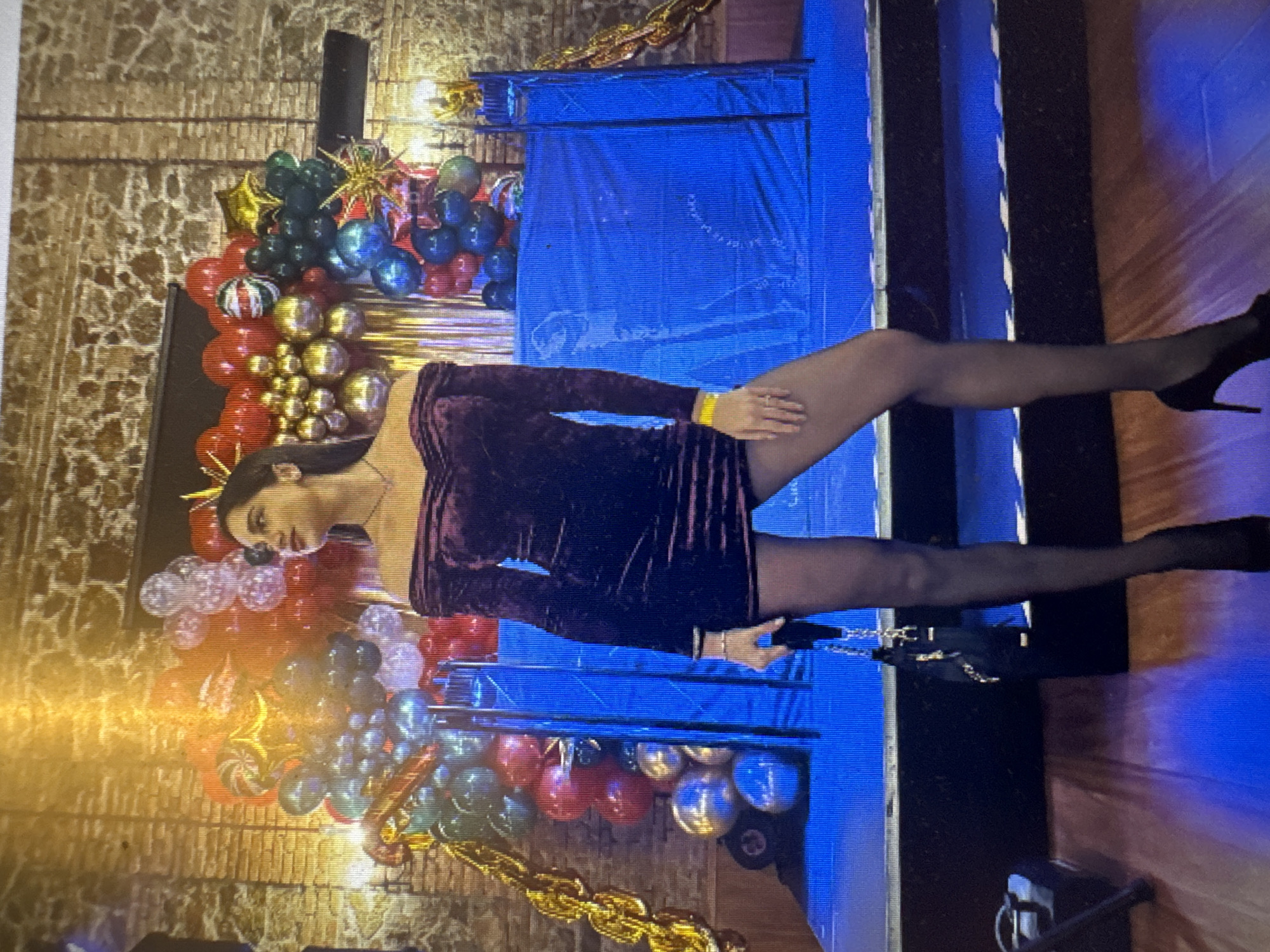 In the city of Murcia, Spain, a woman stands on a raised platform decorated with balloons and gold chain-like decorations. She is wearing a dark, off-the-shoulder velvet dress, black pantyhose, and black high heels. She is holding a small black purse with a chain strap. The background features a brick wall and a blue backdrop.