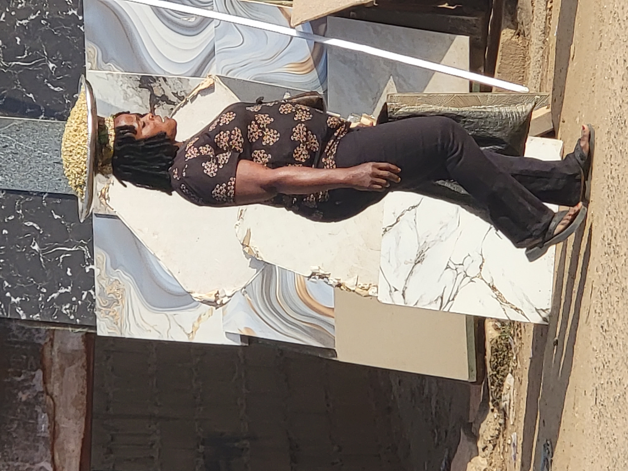 A woman lies on a makeshift bed constructed from stacked ceramic tiles in an outdoor setting in Maiburiji, Nigeria. The woman is lying on her back, wearing a black top with tan floral patterns, black pants, and sandals. A shallow metal dish filled with small yellow objects (possibly grains or seeds) is placed on her head. The tiles are arranged in a patchwork fashion, creating a horizontal surface. The environment appears to be a roadside or commercial area, with sandy soil visible on one side. The scene is bathed in bright daylight.