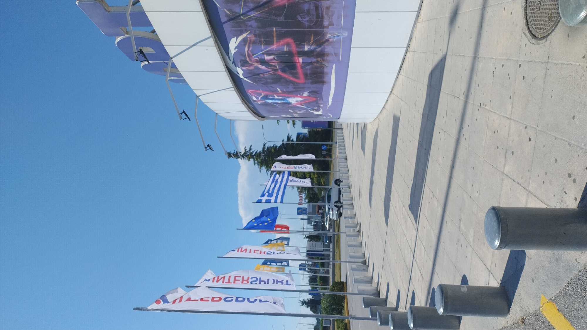 A frontal, medium shot captures a modern building with a curved facade adorned with a large purple and blue mural. The building is situated in Pylaia Municipal Unit, Greece. In front of the building, a row of cylindrical concrete bollards lines a paved walkway. A line of flags, including the European Union and Greek flags, flutters on poles along the walkway. Behind the flags, a white car is visible on a road. The sky above is clear and blue, with a few scattered clouds. The scene is illuminated by bright sunlight, casting long shadows from the bollards and flags onto the pavement.