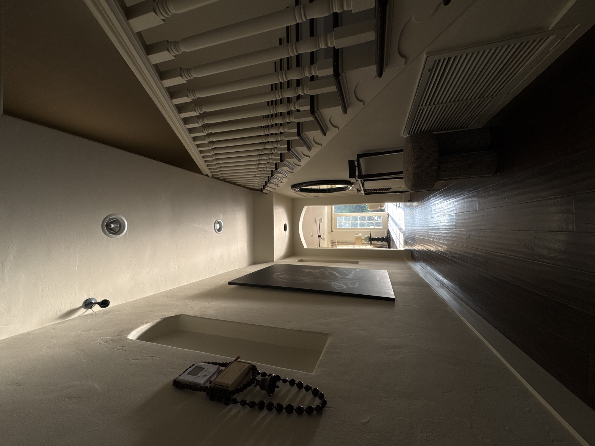 The image displays the interior of a residential hallway or foyer, located in the city of Los Angeles, United States of America. The perspective is low, looking down a long corridor with dark, reflective wooden floorboards towards a brightly lit room at the far end.
The left wall, textured and light-colored, features a wall-mounted spotlight near the ceiling and a recessed arched niche below it. A digital thermostat or control panel is affixed to the wall, from which a string of dark beads with a small metallic bell hangs. Further down this wall, a large, dark, rectangular panel or piece of art leans against it.
On the right side of the frame, a staircase ascends, characterized by numerous light-colored balusters, a matching handrail, and dark stair treads. Beneath the staircase, a large, white rectangular air return vent is visible. A small, cylindrical, textured upholstered stool sits next to the vent. Further along this wall, under the stairs, a dark console table is positioned with a circular mirror mounted above it. The ceiling is light-colored and fitted with recessed lighting fixtures; a contrasting darker, brown-colored section of the ceiling is present above the staircase.
The hallway terminates in an arched doorway leading to another room. Through this doorway, strong natural light floods in from large, multi-paned glass doors or windows, illuminating a ceiling fan and a table with a lamp. No individuals, actions, or direct interactions are depicted in the scene.