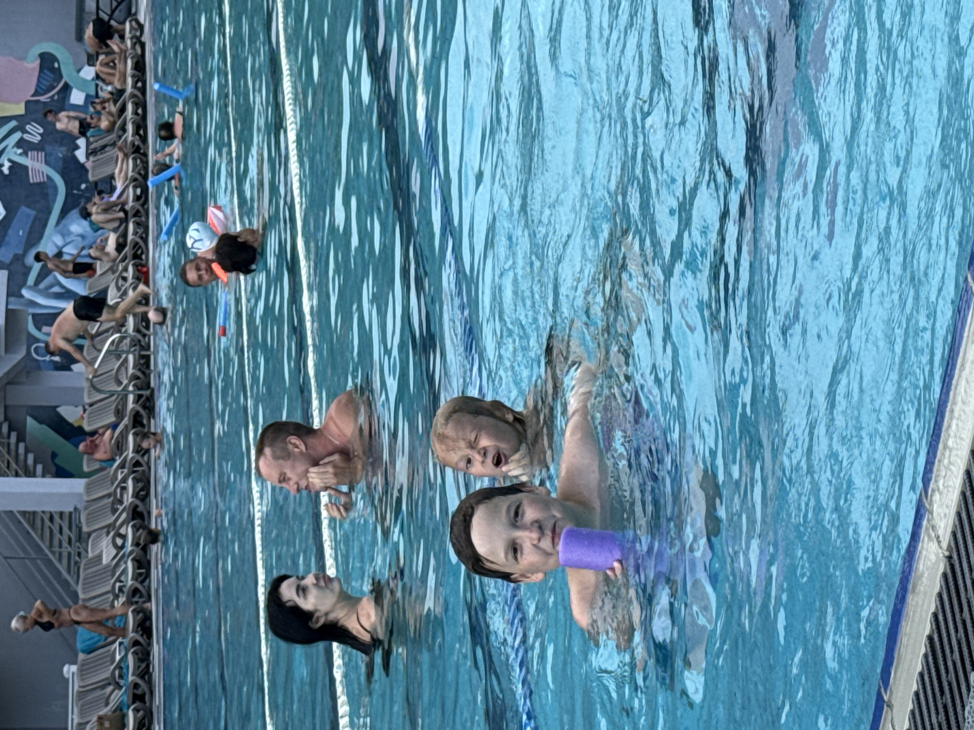 A medium shot captures a swimming pool area with several individuals enjoying the water and surrounding lounge chairs. In the foreground, two young boys are in the pool. The boy on the left, facing the camera, holds a purple pool noodle and smiles. Behind him, another boy with light hair makes a face while touching his cheek.
Further back, a man and a woman are in the pool, appearing to be in conversation. The man has his hand on his chin, and the woman looks towards him. In the background, several people are relaxing on lounge chairs on the poolside, which is adjacent to a building with a mural on its outer wall. Some individuals are visible in the water further out, with pool noodles scattered throughout the pool. The water is a clear blue, and the overall scene depicts a leisurely day at a public pool. The location is identified as the Massandrivskyi settlement council in Ukraine.