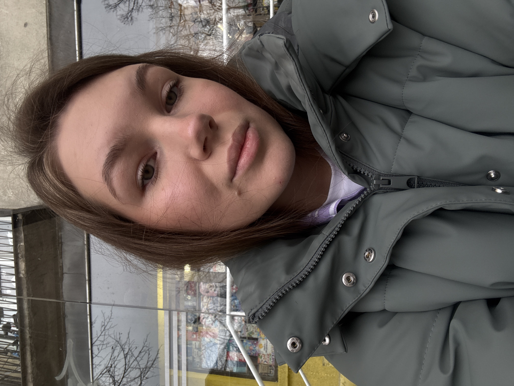 A young woman takes a selfie outdoors, likely in the city of Vozdovac Urban Municipality, Serbia. She is wearing a grey puffer jacket with the zipper partially undone and silver-colored snap buttons. Her brown hair is parted to one side. A glass window is visible behind her, reflecting a tree and what appears to be an interior space with shelves. A white railing is also visible behind her.