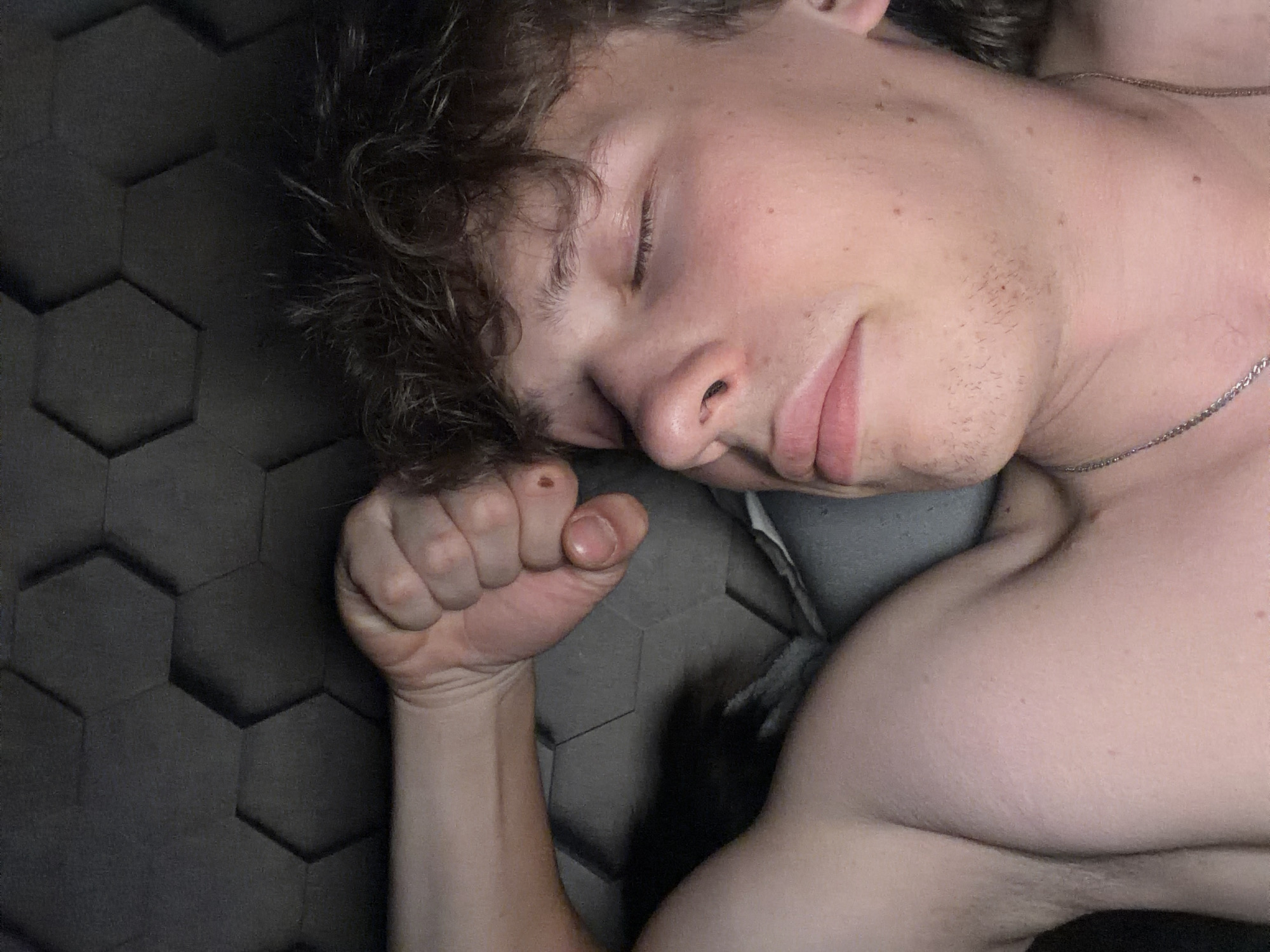 The image is a close-up, overhead shot of a young person lying down with their eyes closed, appearing to be asleep or resting. They have curly brown hair and are shirtless, wearing a silver chain necklace. Their right arm is bent, with their fist clenched and resting near their head. The background consists of a dark gray, hexagonal textured surface, which could be acoustic foam or a decorative wall panel. There is no indication of the specific location within Nizhny Novgorod, Russia. The lighting is dim, casting soft shadows.