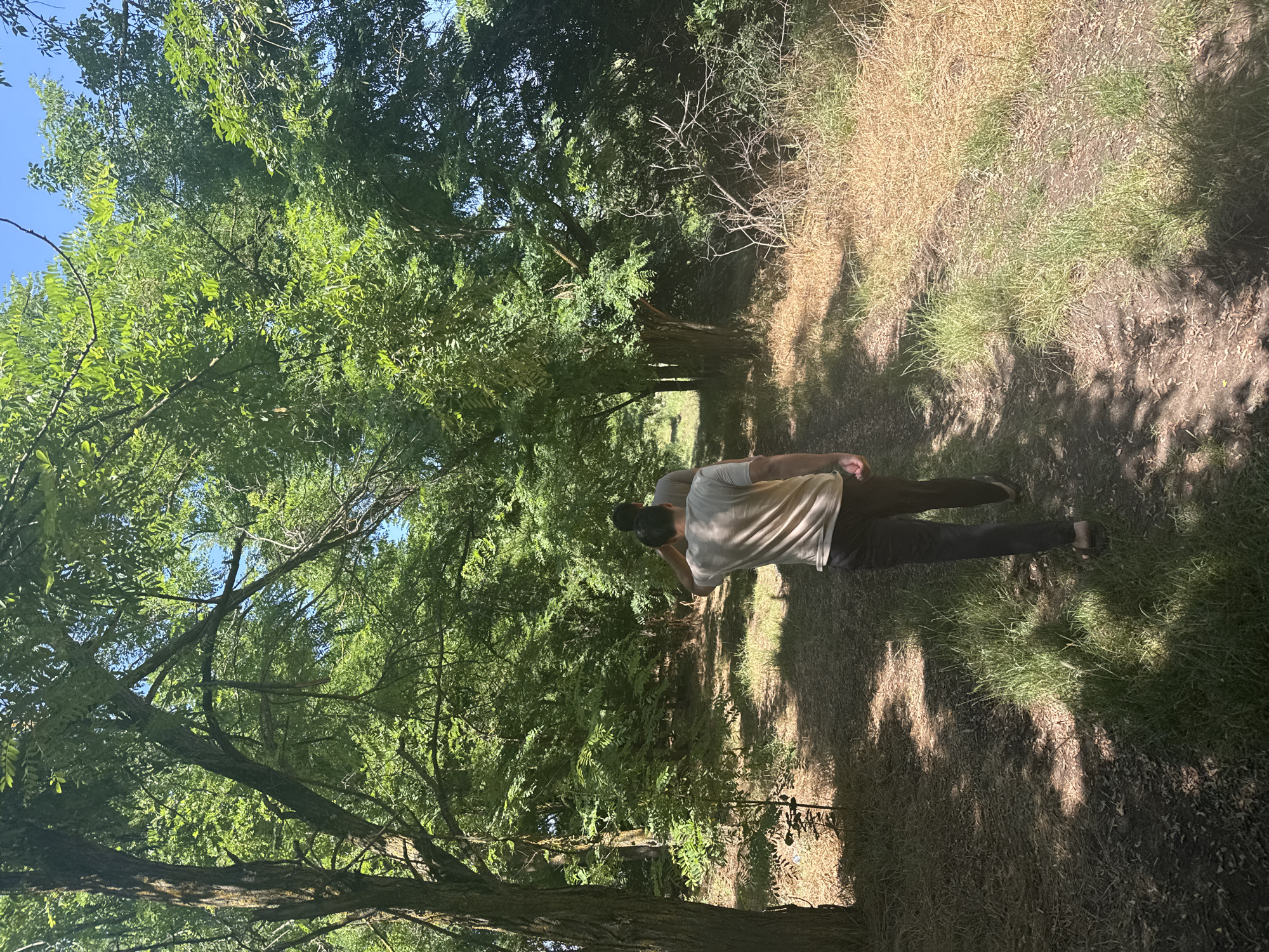 Two individuals are walking on a dirt path through a densely wooded area with a clear blue sky overhead. The person in the foreground, seen from the back, is wearing a light-colored t-shirt and dark pants. They appear to be walking away from the camera. Behind them, a second individual, also seen from the back, is walking in the same direction. The path is bordered by lush green trees and dry, brown grass. The sunlight filters through the leaves, casting dappled shadows on the ground. The location is identified as Zarevo, Russia.