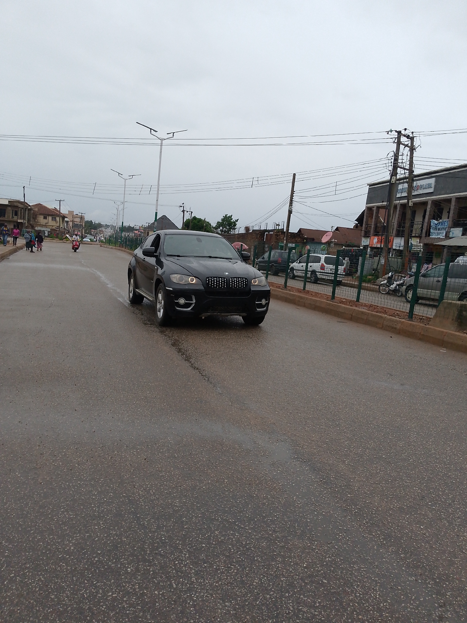This media file presents a street scene in Bukuru, Nigeria, captured on an overcast day. The asphalt road surface appears wet, suggesting recent rain or damp conditions.
The main subject is a black BMW X6 SUV, prominently featured in the foreground, driving towards the viewer. Its headlights, including the distinctive circular daytime running lights, are illuminated.
On the left side of the road, several people are walking on a dirt shoulder, and further in the distance, a person is riding a moped. This side is lined with various multi-story buildings, some appearing residential and others commercial, showcasing a mix of architectural styles.
The right side of the road features a green metal fence separating the main thoroughfare from a row of parked vehicles and additional buildings. A large, multi-story structure with a grey facade is visible here. Numerous utility poles supporting a complex network of power lines are prominent on this side. A banner hanging from one of the buildings partially reads "...ONES / FAJA VENTUR / MEMORIES", and another sign above it faintly shows "PLAY ZONE".
The sky is uniformly grey, indicating heavy cloud cover. Modern streetlights with dual luminaires extend into the distance, lining the road. The scene depicts a typical busy thoroughfare in an urban or semi-urban setting, combining daily life with infrastructure.