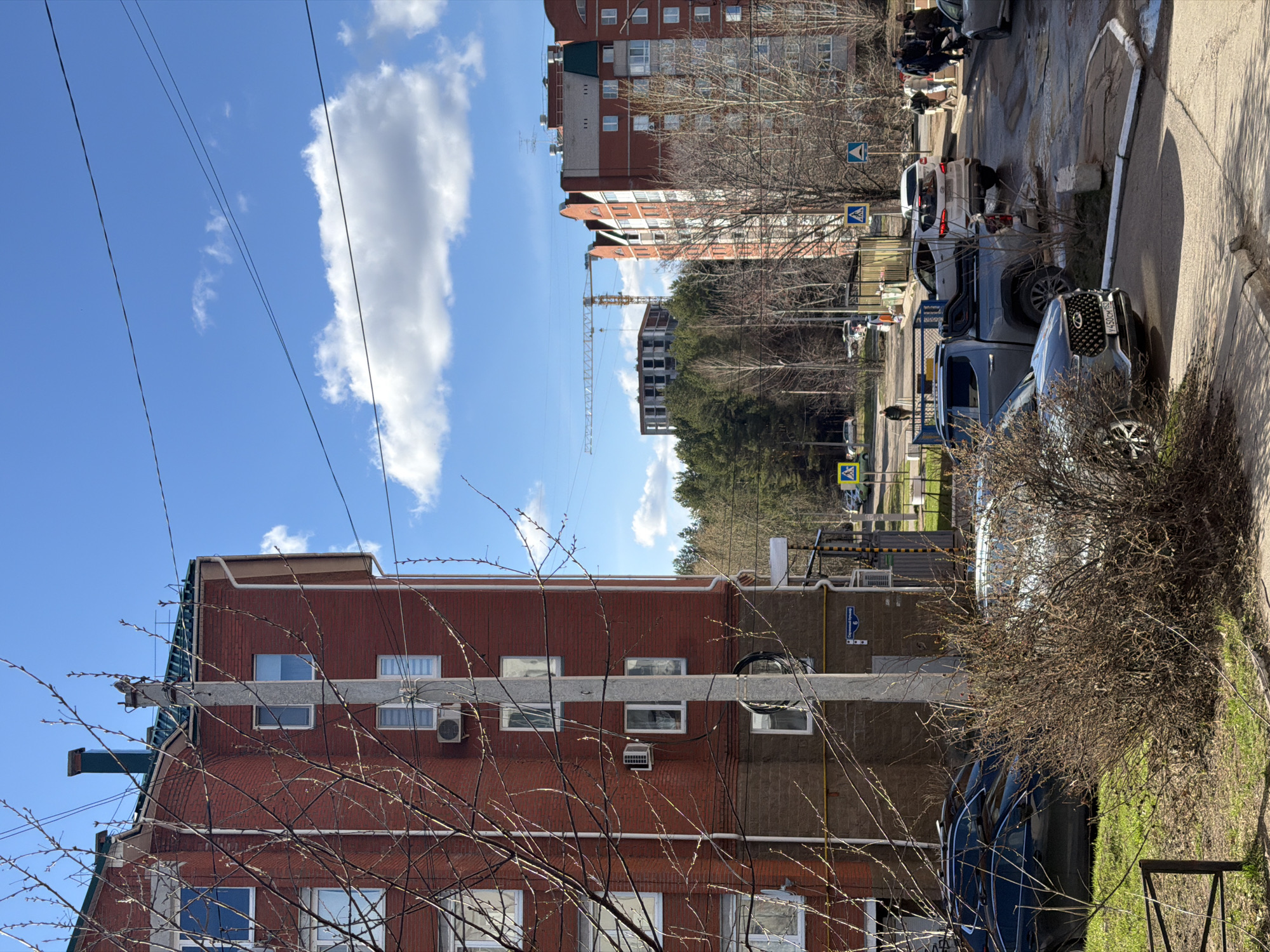 The image depicts a street scene in Chaikovsky, Russia. In the foreground is a red brick building with multiple windows and air conditioning units. A utility pole runs vertically along the building, with bare branches obscuring part of the view. Several cars are parked along the street. In the background are taller apartment buildings and a construction crane. A pedestrian crosswalk sign is visible. The sky is blue with some clouds. People are walking in the background.