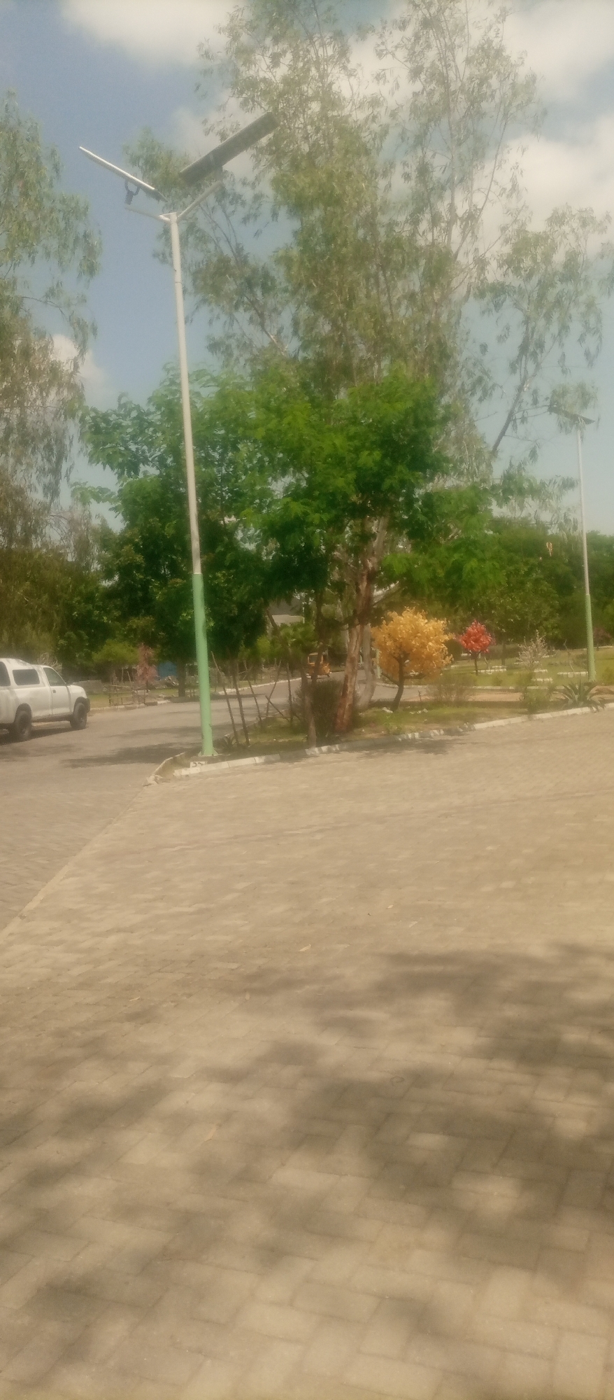 The image shows a paved area with trees and a solar-powered street light in Kuson, Nigeria. A white vehicle is parked on the left side of the paved area. There are several trees with green foliage, and one tree with yellow and red leaves. The sky is cloudy. The ground is paved with interlocking bricks. In the background, there are more trees and a fence. A small, three-wheeled vehicle is visible in the distance. The scene appears to be an outdoor public space.