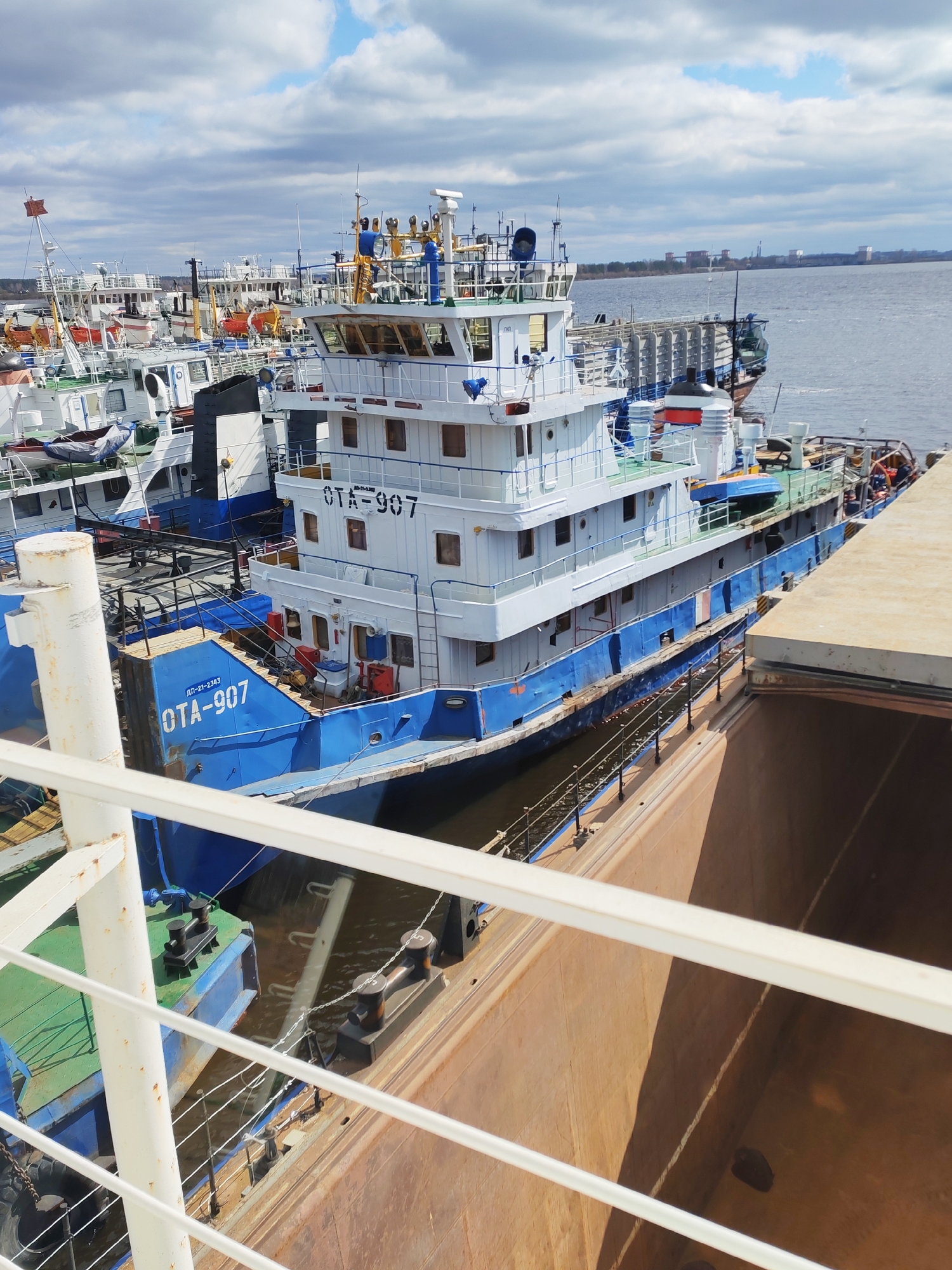 The image shows a view from a ship, possibly on the Volga River, in Rybinsk, Russia. Multiple water vessels are docked, with a prominent blue and white tugboat labeled "OTA-907" taking center stage. The surrounding environment includes other docked boats, the river, and distant buildings on the opposite bank, beneath a sky partially covered by clouds. The foreground view from the ship captures details of its railings and a portion of a large brown structure.