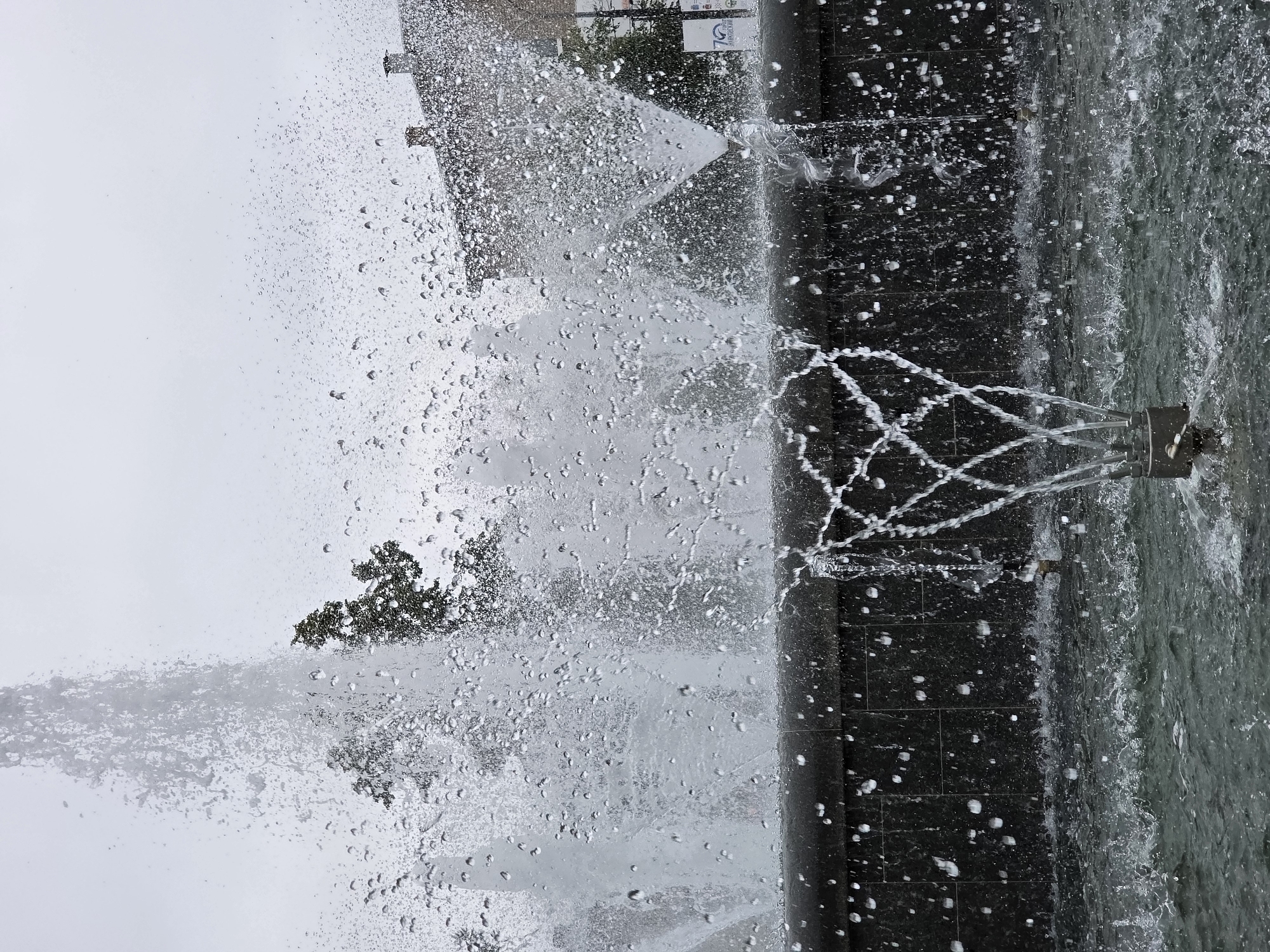 The image shows a fountain in the city of Chaikovsky, Russia, on an overcast day. Water is spraying upwards from multiple nozzles, creating a misty spray that obscures the background. The fountain has a dark, tiled basin, and the water collects in a pool at the bottom. In the background, trees and buildings are visible, but they are out of focus due to the water spray and the overall lighting conditions. There are no people visible in the image. The overall impression is one of movement and motion, with the water creating a dynamic and somewhat abstract scene.