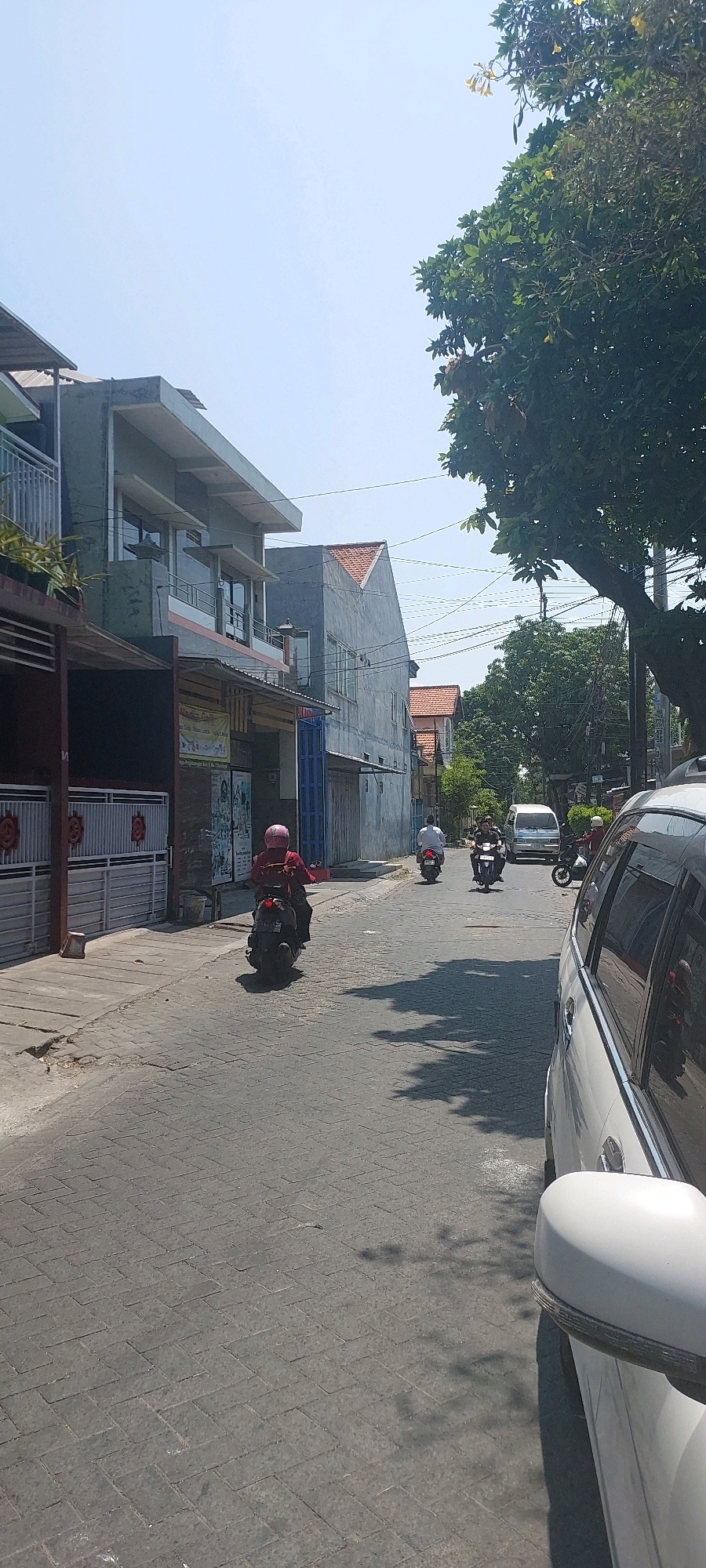 A vertical, medium shot captures a sun-drenched street scene in Surabaya, Indonesia. The asphalt road, paved with interlocking bricks, is lined with multi-story buildings. On the left, a building features a grey facade with balconies and glass windows. Further down the street, other buildings exhibit a similar architectural style, some with red-tiled roofs. A large tree with dense green foliage dominates the right side of the frame, casting dappled shadows on the road.
Several individuals are present, all on motorcycles. The closest is a person wearing a red shirt and pink helmet, riding a black scooter. Further down, another scooter rider in a white shirt is visible, followed by a white van. A third motorcycle is parked on the right side of the road, with its side visible in the foreground. The sky is clear and blue, suggesting a bright day. The overall impression is of a typical urban street in a tropical climate.