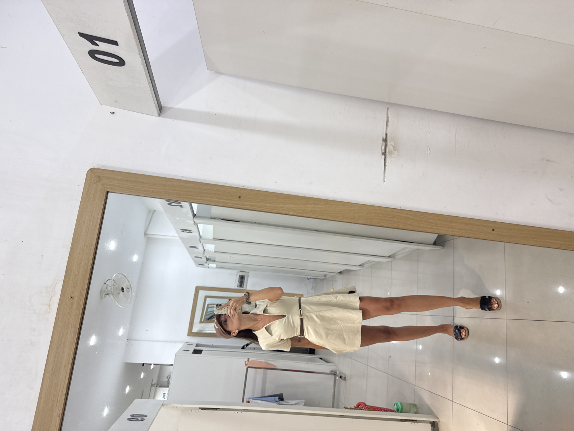 A mirror reflects a woman in a cream dress, positioned in a retail setting. The room includes clothing racks, white storage units, and a fan. The number "01" is visible on a white block near the mirror. The location may be Đà Nẵng, Vietnam.