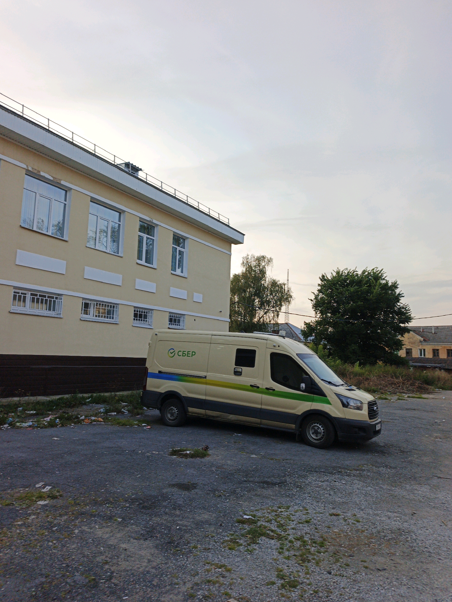 A beige van with a blue, green, and yellow stripe along its side and the green SBER logo is parked on a gravel lot. The van is a Ford Transit model. To the left of the van is a light yellow building with multiple windows. The building has a white trim and a railing along its roofline. The ground is covered in gravel and some patches of grass, with scattered litter near the base of the building. In the background, there are trees and some dilapidated buildings. The sky is overcast. The image was taken in Zlatoust, Russia.