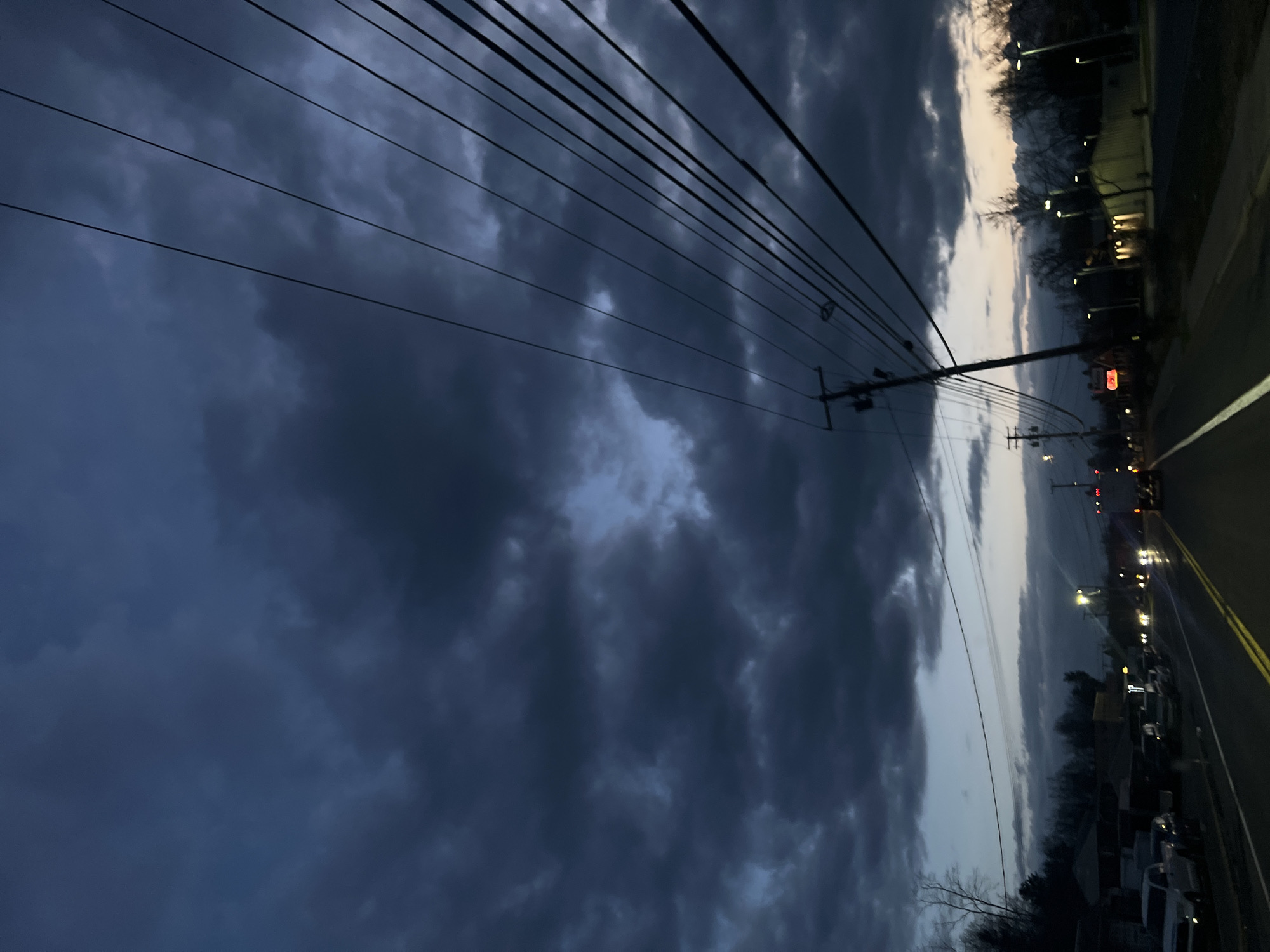 A low-angle, landscape-oriented photograph captures a street scene in the city of Newburgh, United States, under a dramatic, cloudy sky. The time of day appears to be dusk or early evening.
The primary subject is the heavy, overcast sky filled with dark, swirling clouds that dominate the majority of the frame. High-tension power lines run across the top of the image, intersecting with utility poles on the right side.
On the right side of the photograph, a street runs into the distance. The road is marked with yellow lines. Along the roadside are parked cars, buildings, and street lights that cast a soft glow. Red traffic signals can be seen further down the road.
The photograph is slightly tilted, giving a sense of motion or an off-kilter perspective. The overall mood is somber due to the dark clouds and dim lighting.
