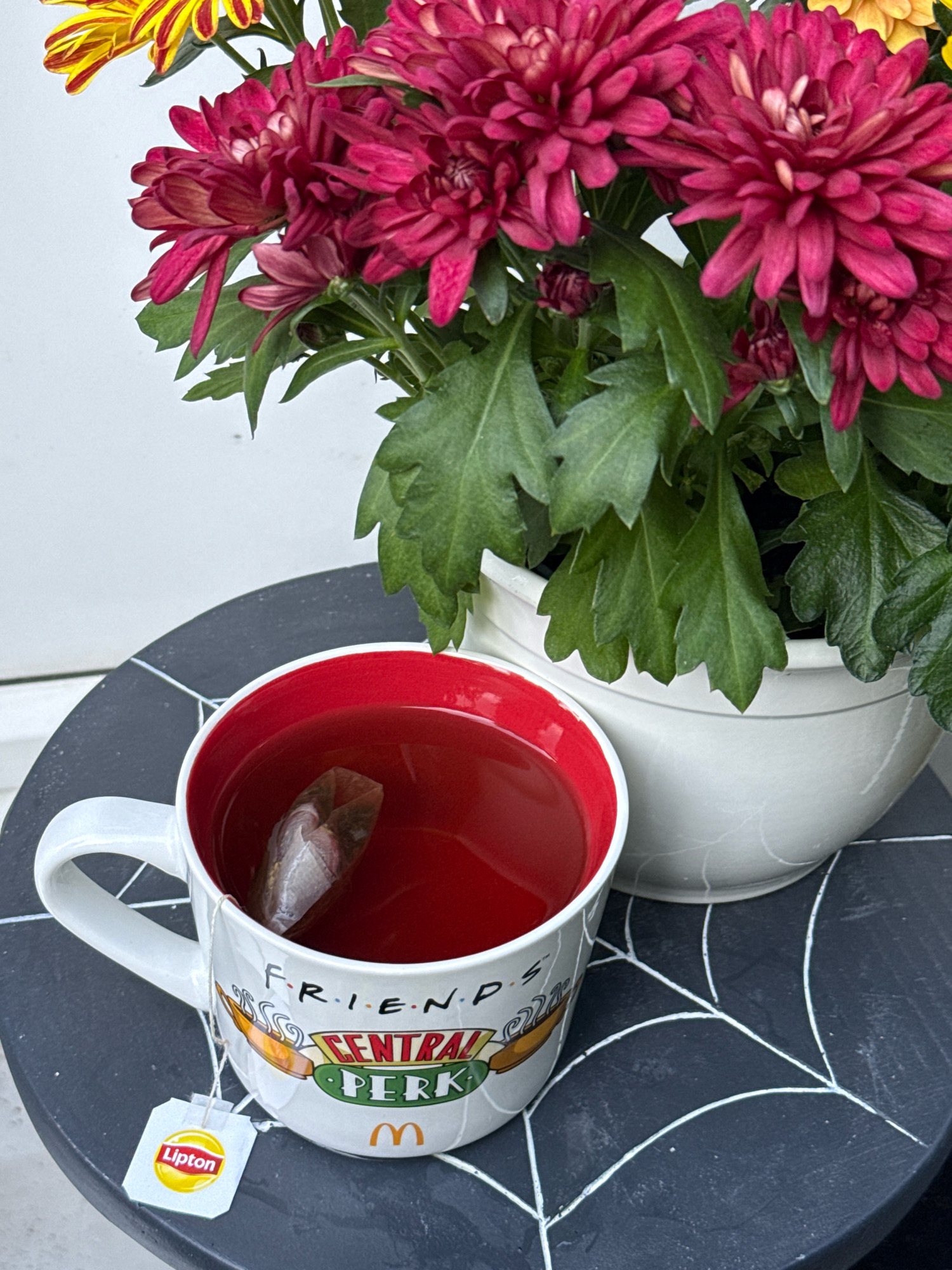 A white mug with a red interior, decorated with the "Friends" Central Perk logo and a McDonald's logo, sits on a black round table with a white spiderweb design. Inside the mug is a reddish-brown liquid, presumably tea, with a tea bag immersed. A Lipton tea bag tag hangs outside the mug. Behind the mug, a white ceramic pot contains a cluster of deep pink chrysanthemums, with hints of yellow flowers visible in the upper left corner. The background is a plain white wall. The scene is set in the city of Bad Sachsa, Germany.