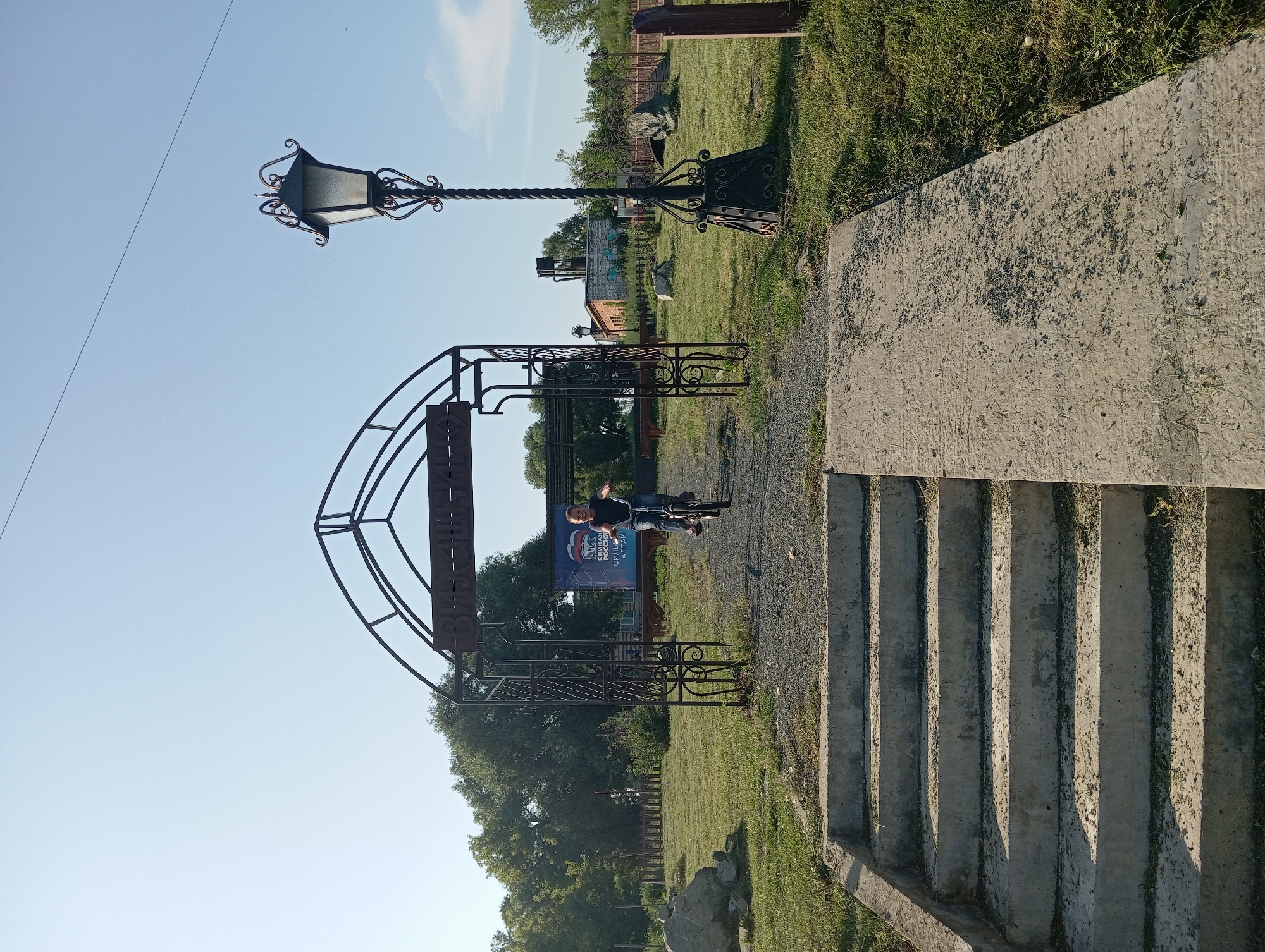The image shows a bright, sunny day in Новокопылово, Russia. In the foreground, there are concrete stairs leading up to a gravel path. To the right of the stairs is a concrete ramp. Beyond the path is an ornate, wrought-iron archway with Cyrillic lettering that appears to say "ВЛАДИМИРСКОЕ". A young person is riding a bicycle through the archway. To the right of the archway is a tall, decorative lamppost. The background features a grassy field, trees, and a brick building. A banner with Russian text and a bear emblem is visible behind the archway. The sky is clear and blue.