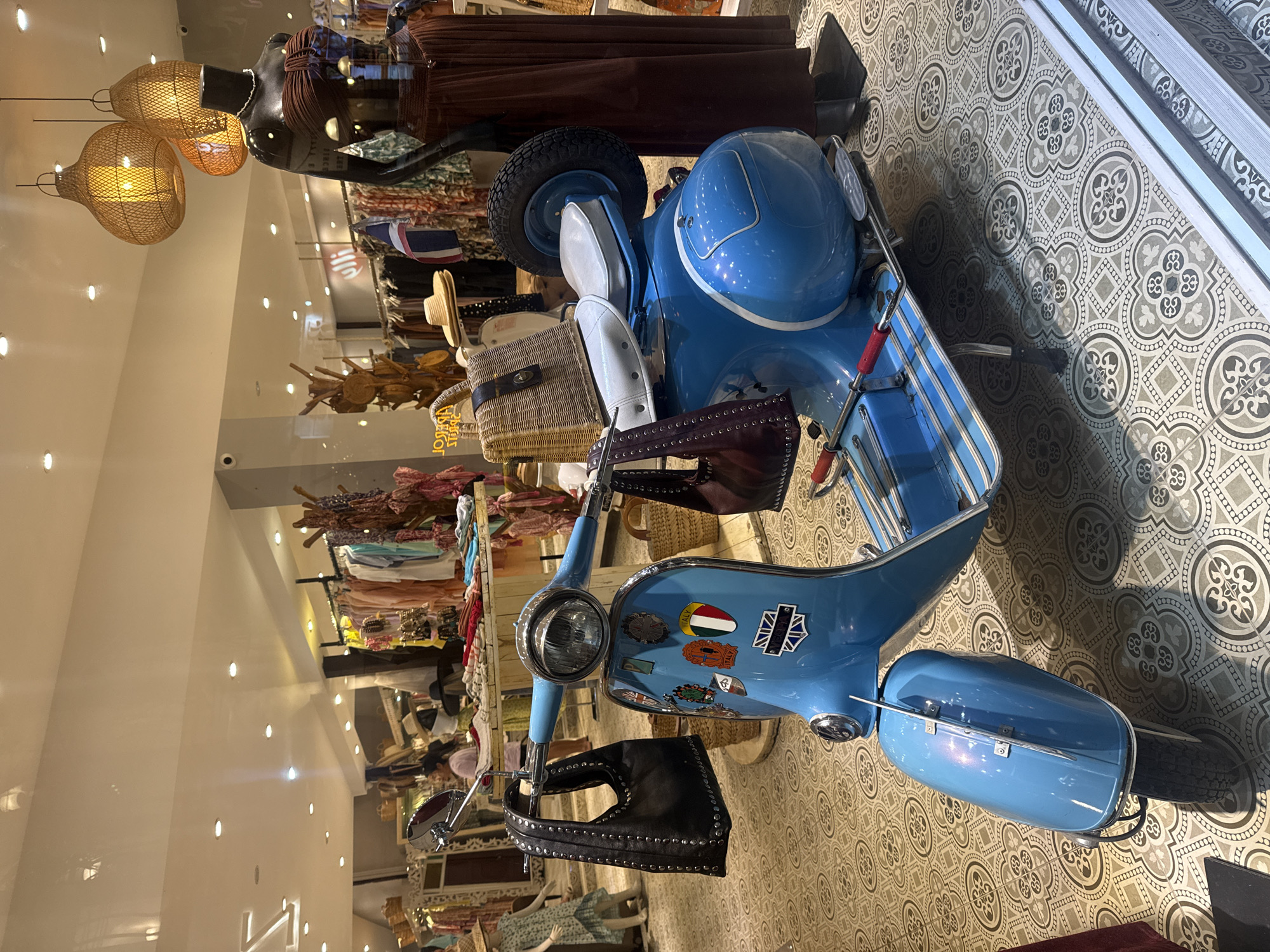 In a retail store in Ko Samui District, Thailand, a blue vintage scooter is displayed prominently on a patterned tiled floor. The scooter is accessorized with two handbags: a black studded bag hanging from the handlebars and a dark red bag with a strap on the seat. A wicker basket is also placed on the seat. Stickers are affixed to the front panel of the scooter, including flags of Italy and the United Kingdom. In the background, clothing racks filled with apparel, mannequins dressed in fashionable clothing, and decorative lamps are visible, suggesting a boutique or clothing store setting. The lighting in the store is warm and inviting.