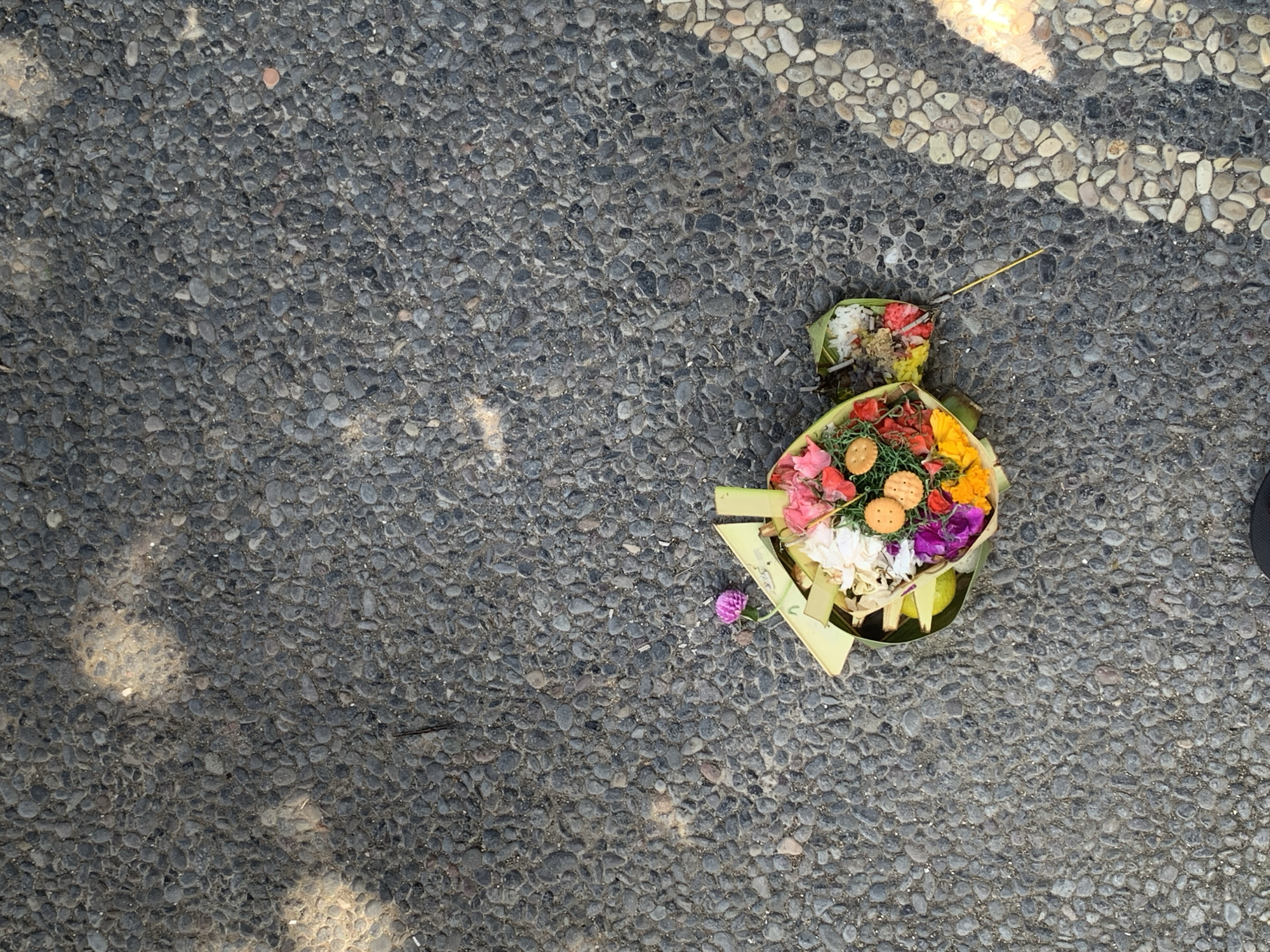 A small offering basket, typically called a canang sari, lies on a pebble-strewn pavement in Sukawati, Indonesia. The basket, woven from banana leaves, contains a colorful arrangement of flowers, rice, and small crackers. Sunlight casts dappled shadows across the ground, highlighting the textures of the stones and the offerings. A single pink flower with a delicate stem stands out from the basket, and a thin yellow stick extends from the side. The scene captures a moment of cultural practice in a public space, suggesting a roadside or pathway in the town.