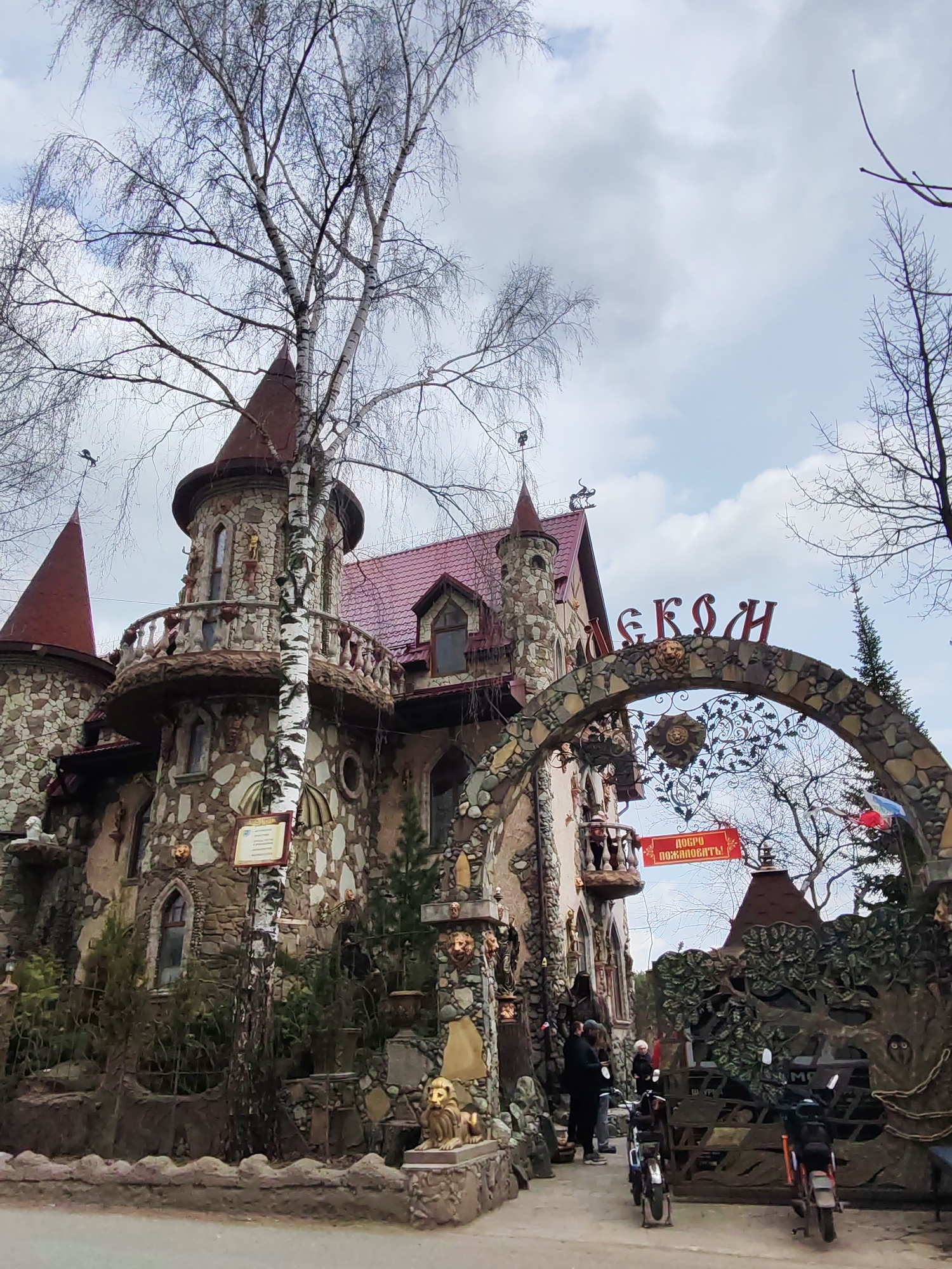 This media file captures the exterior of an elaborately decorated building, resembling a fantasy castle, under an overcast sky. The building is constructed with a mosaic of various stones and features multiple turrets with conical red roofs. Ornate balconies, adorned with sculptures of figures and lion heads, wrap around some of the turrets.
Bare birch trees, their branches devoid of leaves, dominate the foreground and background, suggesting a setting in early spring or late autumn. A small group of people are gathered near the main entrance, beneath a stone archway. They appear to be visitors or patrons, with a few individuals standing by a small fence near the entrance, and one person visible on an upper balcony. Two motorcycles or mopeds are parked on a paved path in front of the archway.
The archway itself is made of stone and features decorative metalwork, including a stylized sun or lion head design. Above the arch, large red letters spell out "СЕКОН" (SEKON). A red banner, hanging from a point near the archway, prominently displays the Russian phrase "ДОБРО ПОЖАЛОВАТЬ!" (DOBRO POZHALOVAT'!), meaning "WELCOME!". Other decorative elements include various sculptures around the building's base, a small informational sign attached to a birch tree, and two flags, one of which is the Russian flag, flying near the entrance. The building's whimsical and eclectic design gives it a fairytale-like atmosphere. The time of day is daytime.