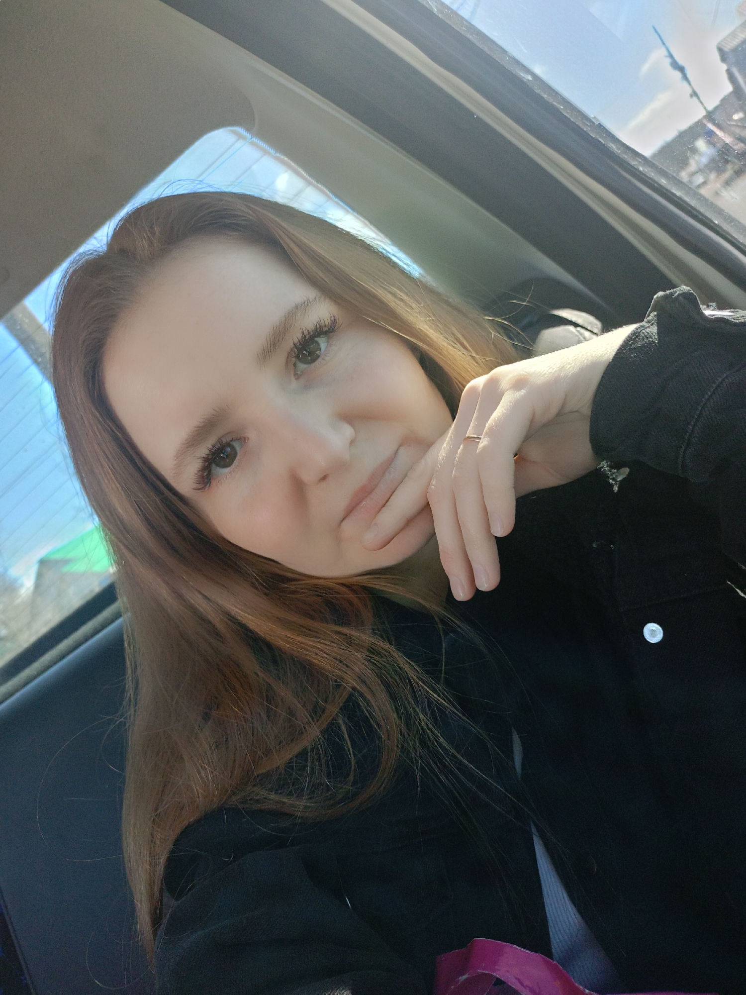 A fair-skinned person with long, light brown hair, wearing a dark jacket, is sitting inside a car. The person is looking towards the camera, resting their chin on their hand, which has a thin ring on one finger. The car window is visible, showing an out-of-focus cityscape with buildings under a bright, sunny sky.
