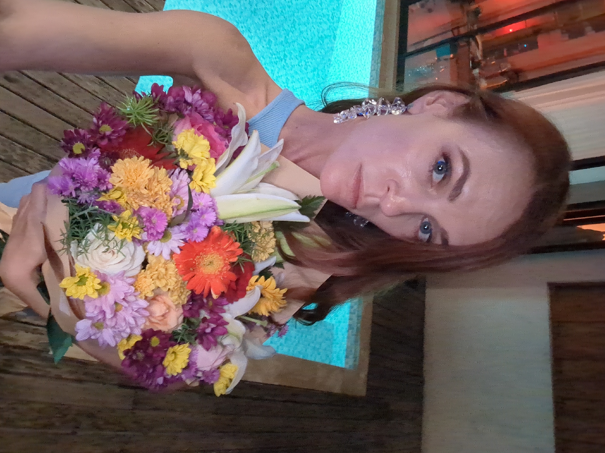A fair-skinned woman with red hair is holding a bouquet of colorful flowers near what appears to be a pool. The bouquet contains lilies, daisies, roses, and other various flowers in shades of red, orange, yellow, white, pink, and purple. She is wearing a light blue dress or top and earrings with crystal-like beads. The setting is likely an outdoor location, possibly a resort or private residence, with wooden decking visible. The woman is in focus, and the background includes a partially visible building. There is no indication of the city of Kediri, Indonesia, in the image.
