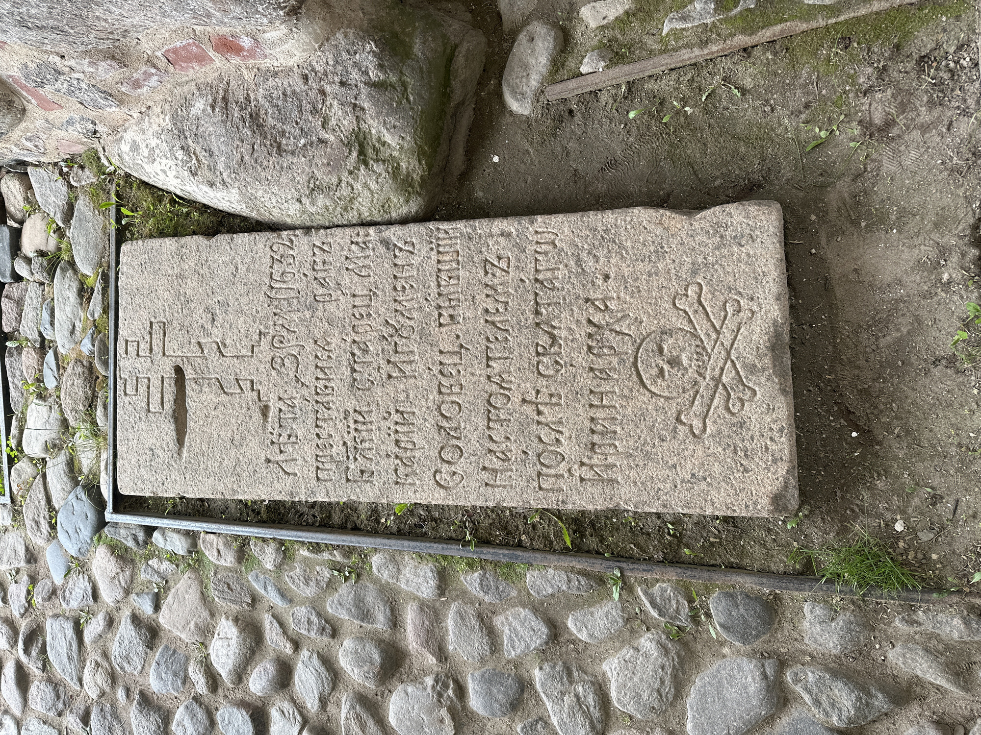 This image shows a weathered stone slab with carved inscriptions and a skull and crossbones motif, positioned on a cobblestone path. The slab is framed by a metal border. The inscription appears to be in Cyrillic script. The setting suggests an outdoor historical or memorial site, possibly a cemetery or religious complex. Given the context of the "Solovetskiy Stone" from Solovetskiy, Russia, this is likely a grave marker or memorial to victims of repression. The stones and surrounding earth show signs of age and exposure to the elements, with some moss and grass growing in crevices. A large, rough-hewn stone and a section of brick wall are visible to the right of the slab, further indicating an ancient or historical location.