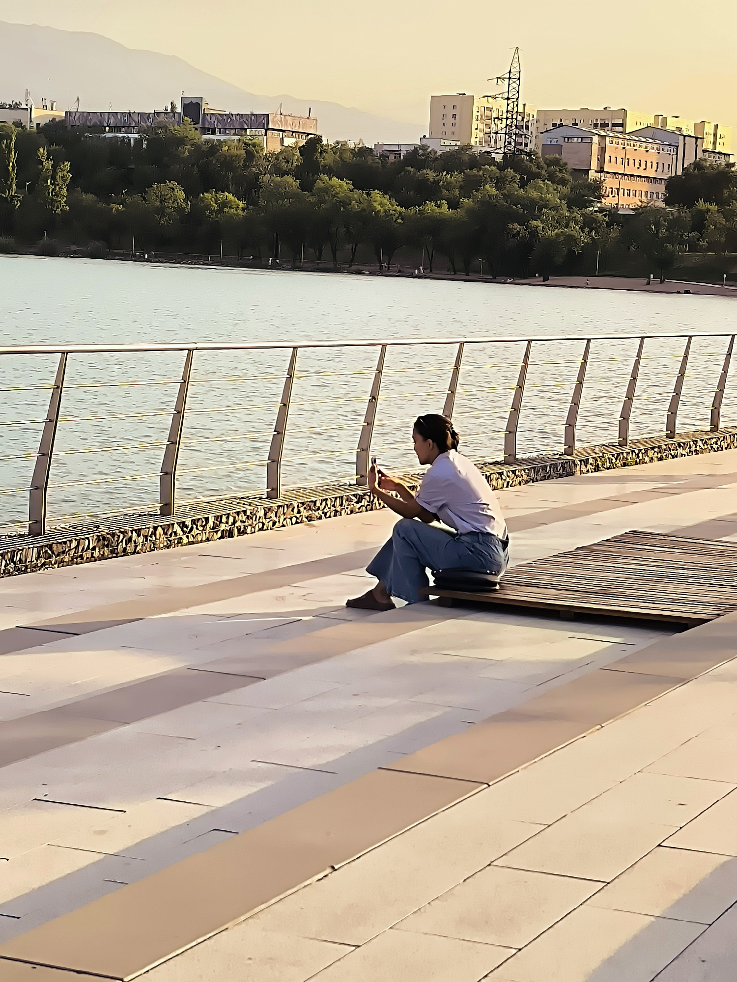 A person sits on a wooden bench on a paved promenade, facing away from the camera, looking at a smartphone. The person is wearing a light-colored short-sleeved shirt and blue pants, with their hair pulled back. The promenade is made of large, light-colored rectangular tiles with darker rectangular accents. A metal railing with vertical supports separates the promenade from a body of water. Beyond the water, there is a tree-lined embankment with several buildings and a distant mountain range. The scene is lit by the warm glow of what appears to be sunset. This location is likely the embankment of the First President's Park in Almaty, Kazakhstan, which features a promenade overlooking a lake and offers views of the city and the surrounding mountains.