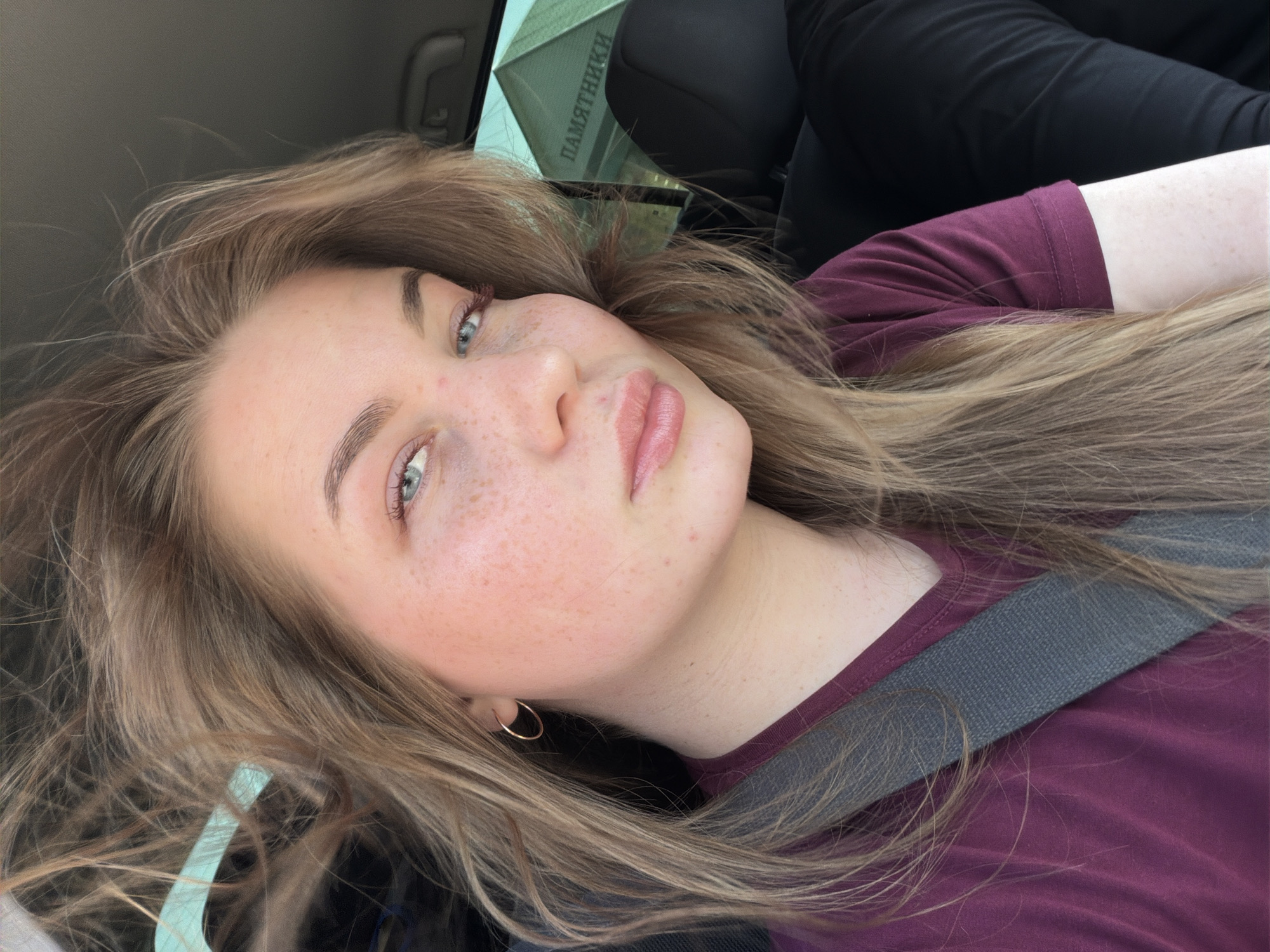 A young woman with light brown hair, freckles, and blue eyes is visible in a close-up, angled selfie. She is wearing a dark purple t-shirt and a seatbelt. An earring is visible in her left ear. Through the car window, the word "ПАМЯТНИКИ" (Pamyatniki), meaning "Monuments," is readable, indicating a location in Russia where funeral monuments are sold. The overall impression is that the woman is a passenger in a car in or near Chaikovsky, Russia.