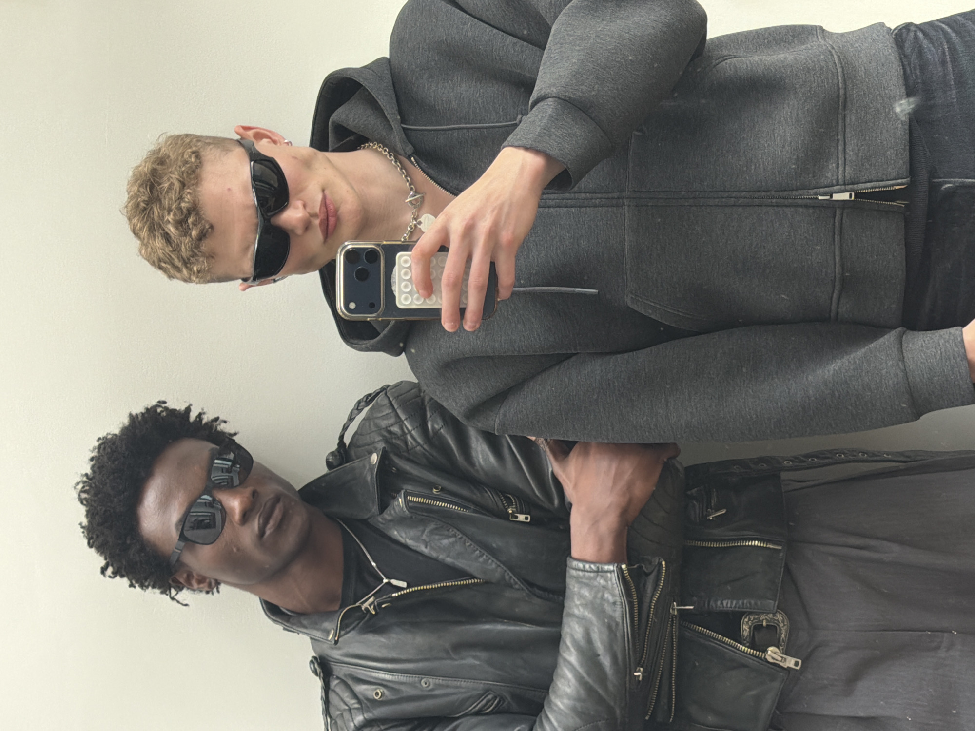 Two people, one fair-skinned with short curly blonde hair, and the other dark-skinned with short curly black hair, are posing for a selfie. Both are wearing sunglasses and facing the camera. The fair-skinned person is wearing a grey hooded sweatshirt and a silver chain necklace with pendants. They are holding a smartphone with a white pop socket on the back. The dark-skinned person is wearing a black leather jacket. The background is a plain white wall. The location appears to be indoors, possibly a studio or an apartment. The image does not provide enough information to confidently ascertain the scene to be in the city of London, United Kingdom.