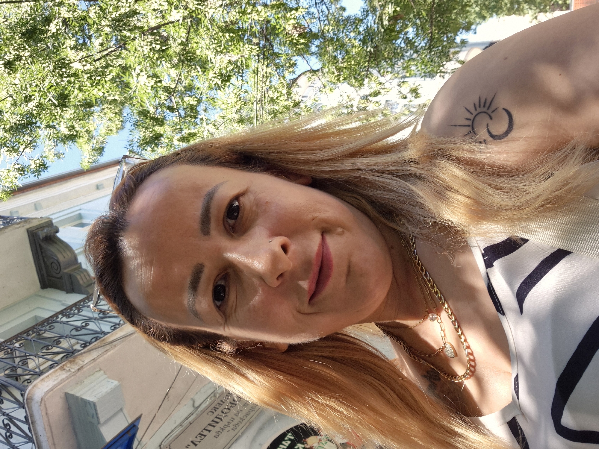 In a close-up, low-angle outdoor shot, a woman with light brown hair is looking at the camera. She is smiling, and her face is illuminated by sunlight. She is wearing a white top with black stripes and several gold necklaces. A tattoo of a sun and moon is visible on her left shoulder. Behind her, a building with ornate balconies and lush green trees are visible against a bright blue sky. The overall scene suggests a sunny day in an urban environment.