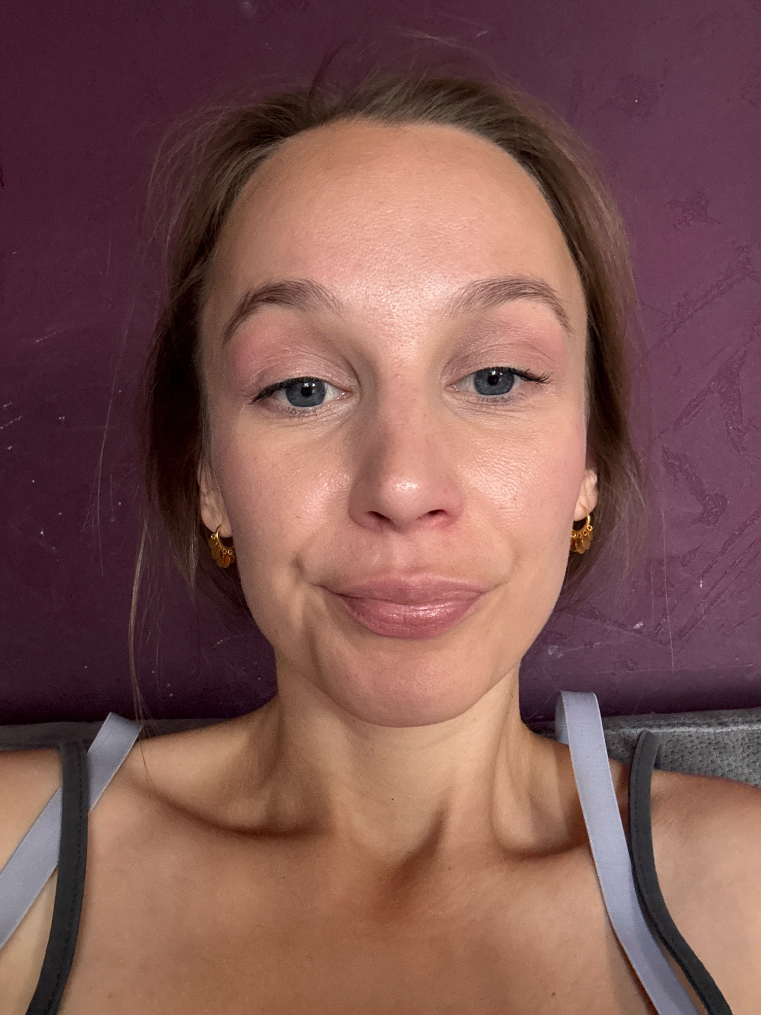 The image is a close-up, head-and-shoulders shot of a young woman with fair skin and light brown hair, taken indoors. She is looking directly at the camera with a slight smile. Her makeup appears natural, with subtle eyeshadow and lip gloss. She is wearing a grey sports bra with light purple straps. The background is a textured purple wall. The lighting suggests an indoor setting, possibly a home. There is no identifiable information to place this image in Überherrn, Germany.