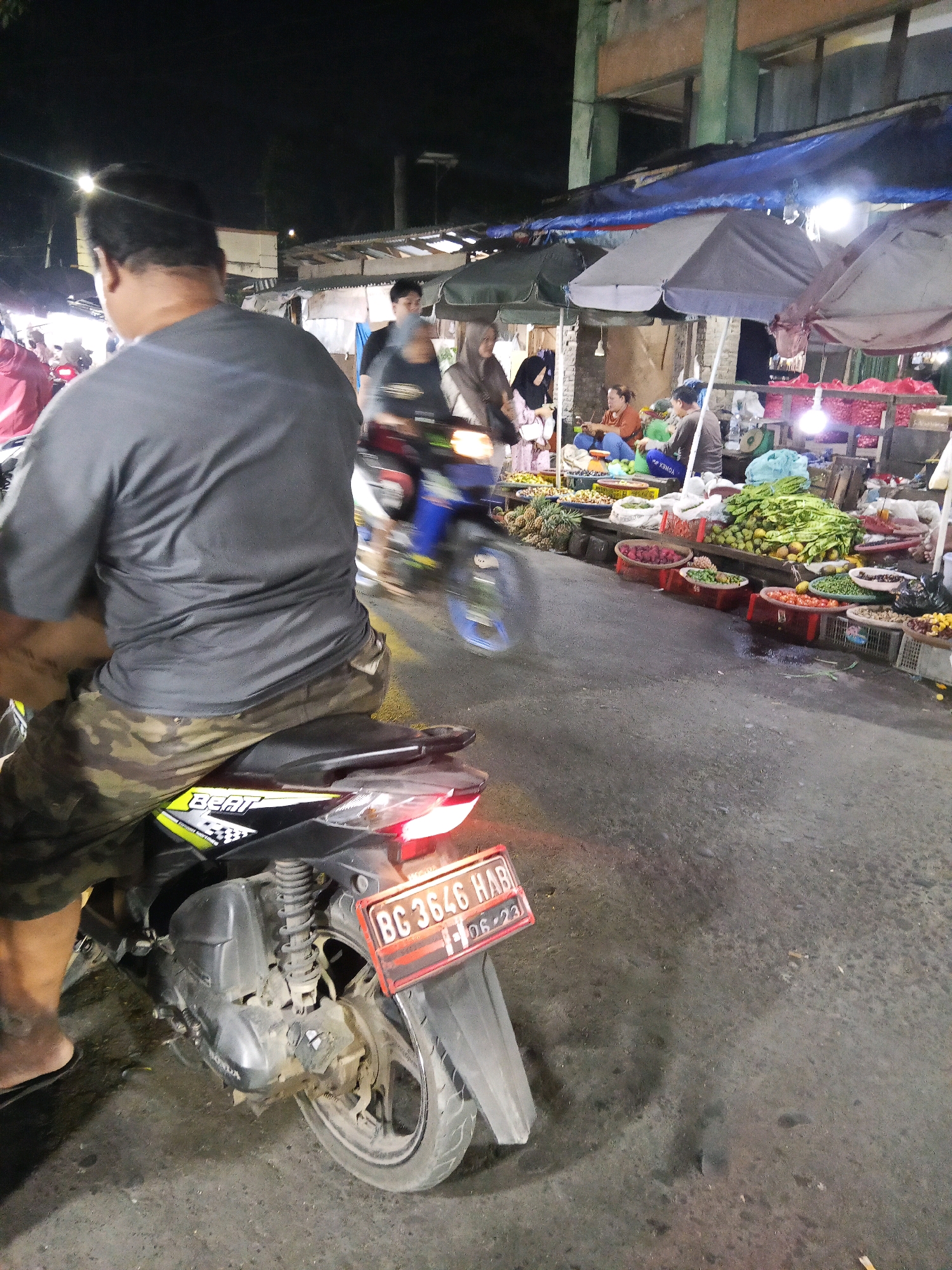 The image shows a busy street market scene at night in Lubuklinggau, Indonesia. In the foreground, a man wearing a grey t-shirt and camouflage shorts is seated on a black and yellow motorcycle. The motorcycle has a license plate that reads "BG 3646 HAB" and "06-23". The man's back is to the camera, and his head is turned to the left. The motorcycle is parked on a paved road that appears wet, with reflections of lights.
In the background, several market stalls are visible, covered by awnings and umbrellas. People are browsing and shopping among the stalls, which are laden with a variety of fresh produce, including pineapples, green vegetables, red fruits, and tomatoes. Bright lights illuminate the market, creating a lively and bustling atmosphere. The overall impression is one of a typical night market in a city in Indonesia.