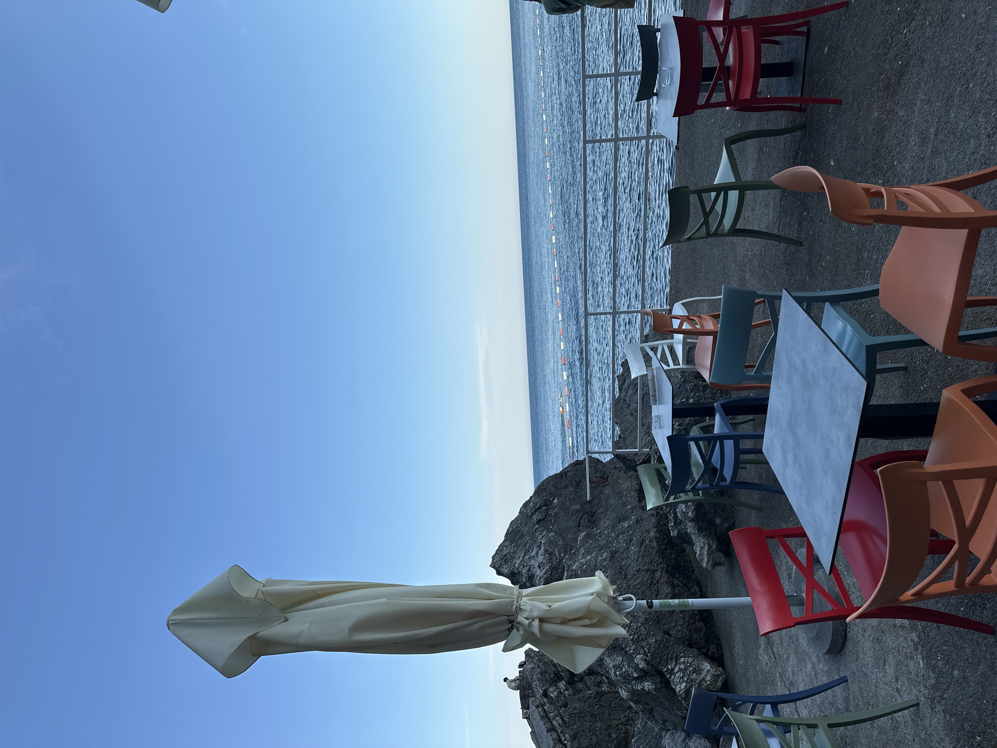The scene depicts an outdoor restaurant or cafe located in Budva, Montenegro, situated on a rocky outcrop overlooking the Adriatic Sea. The restaurant furniture includes colorful plastic chairs (red, orange, green, white, and blue) and a dark-colored rectangular table. A closed white beach umbrella is positioned near the rocks, presumably for shade. The sea is visible in the background, with a line of buoys marking a swimming area. The sky is clear and blue, suggesting a sunny day. No people are visible in the frame.