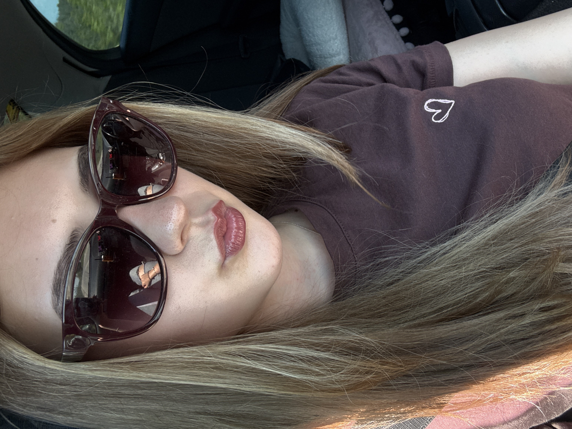 A woman with blond hair is lying down in a car. She is wearing sunglasses and a brown shirt with a small white heart on the chest. The car interior is visible, including parts of the dashboard, seat, and a headrest. The reflection in her sunglasses shows details of a building and other objects. There is no specific information available to place the location in Levski, Bulgaria.