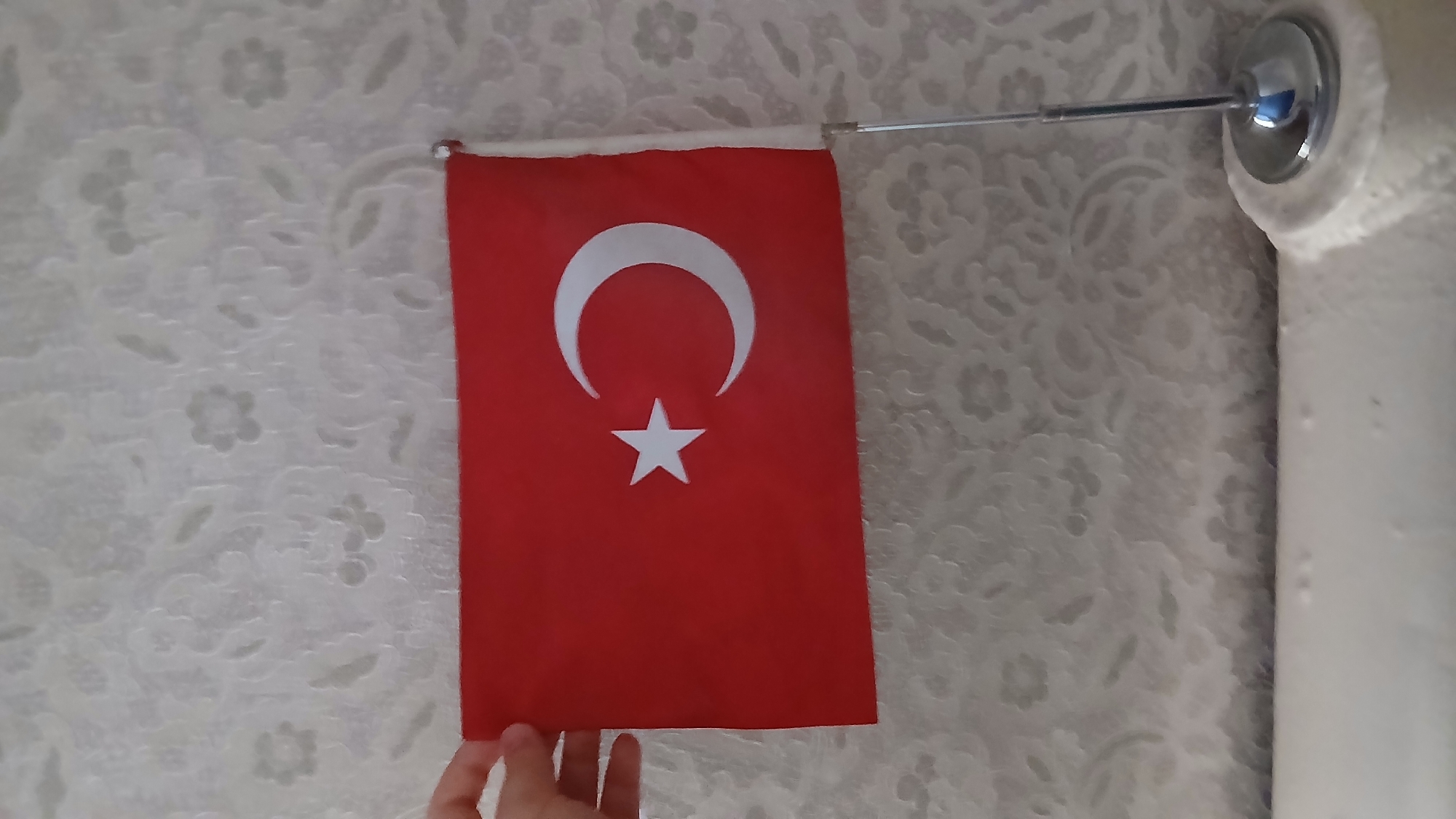 A Turkish flag is held by a hand against a patterned wallpaper background. The flag is red with a white crescent moon and star. A silver flagpole attached to the flag rests on a white surface at the bottom of the image, with a silver base. The overall scene appears to be indoors. The image does not provide any information about the city of Stepnoye, Kyrgyzstan.