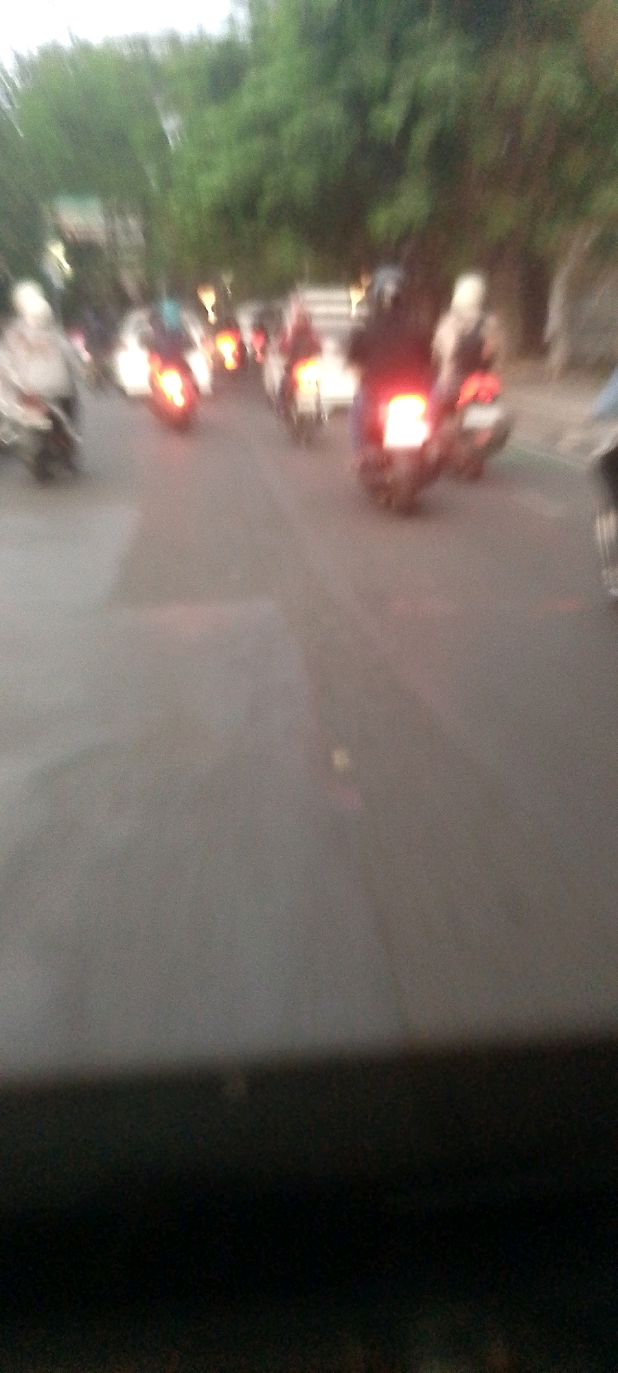 The image is a blurry, low-light photograph taken from a moving vehicle, likely at dusk or dawn, in a city. Multiple motorcycles with illuminated taillights are visible on a paved road, suggesting traffic. Trees line the background, indicating an urban environment. The overall impression is one of motion and indistinct forms. The specific location in Bandung City, Indonesia, cannot be determined from the visual information provided.